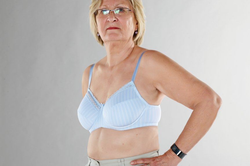 An older woman wearing a blue bra and glasses is standing with her hands on her hips.