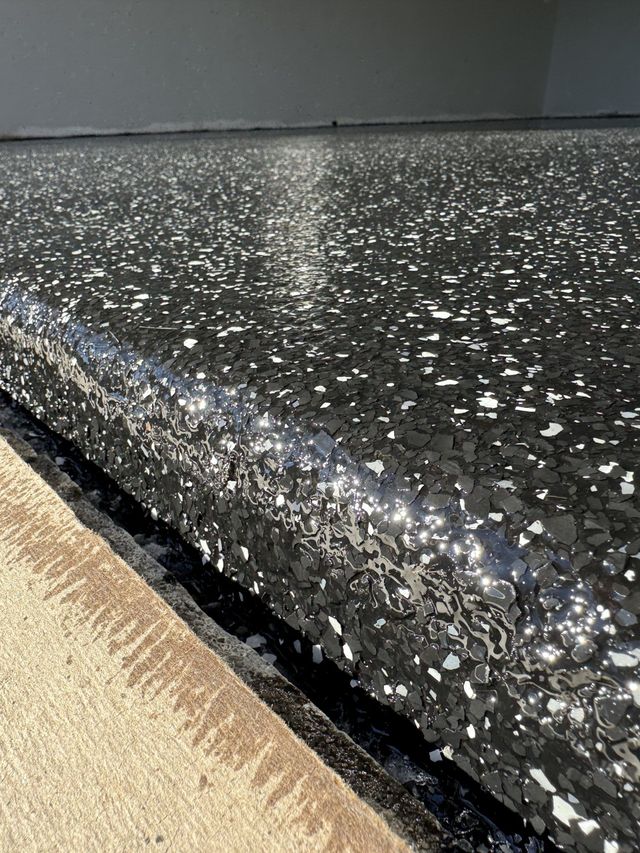 Black glittery textured curb edge beside a beige walkway and gray wall.