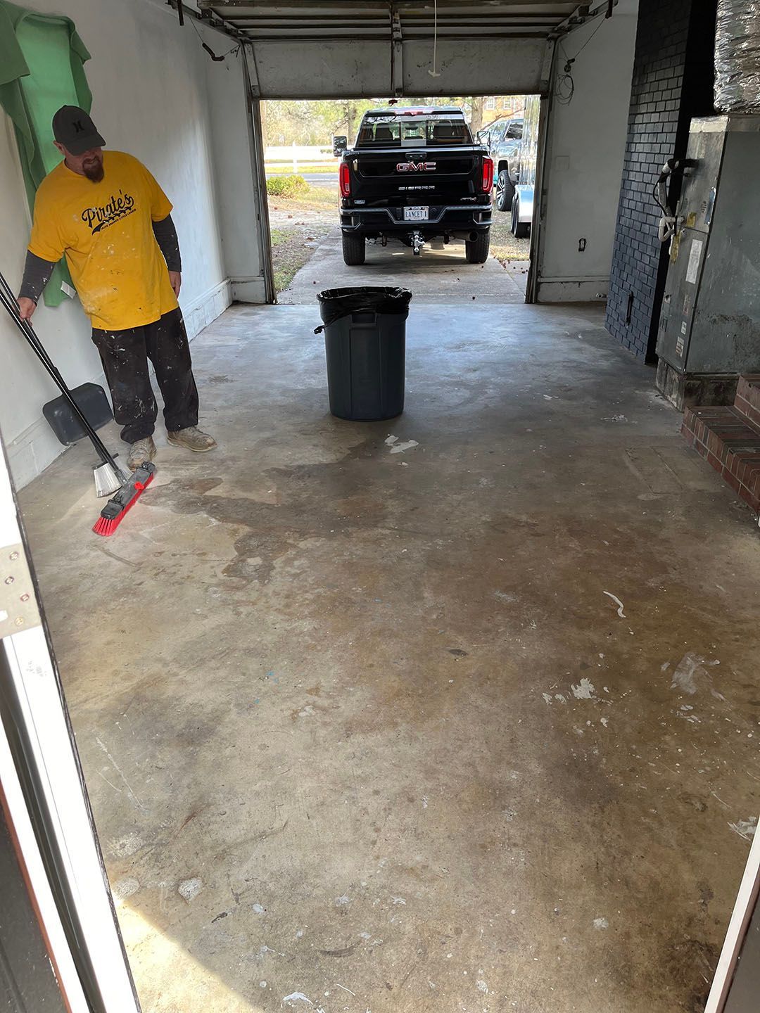 Man sweeping a garage floor near a trash bin, with a black truck parked at the open door.