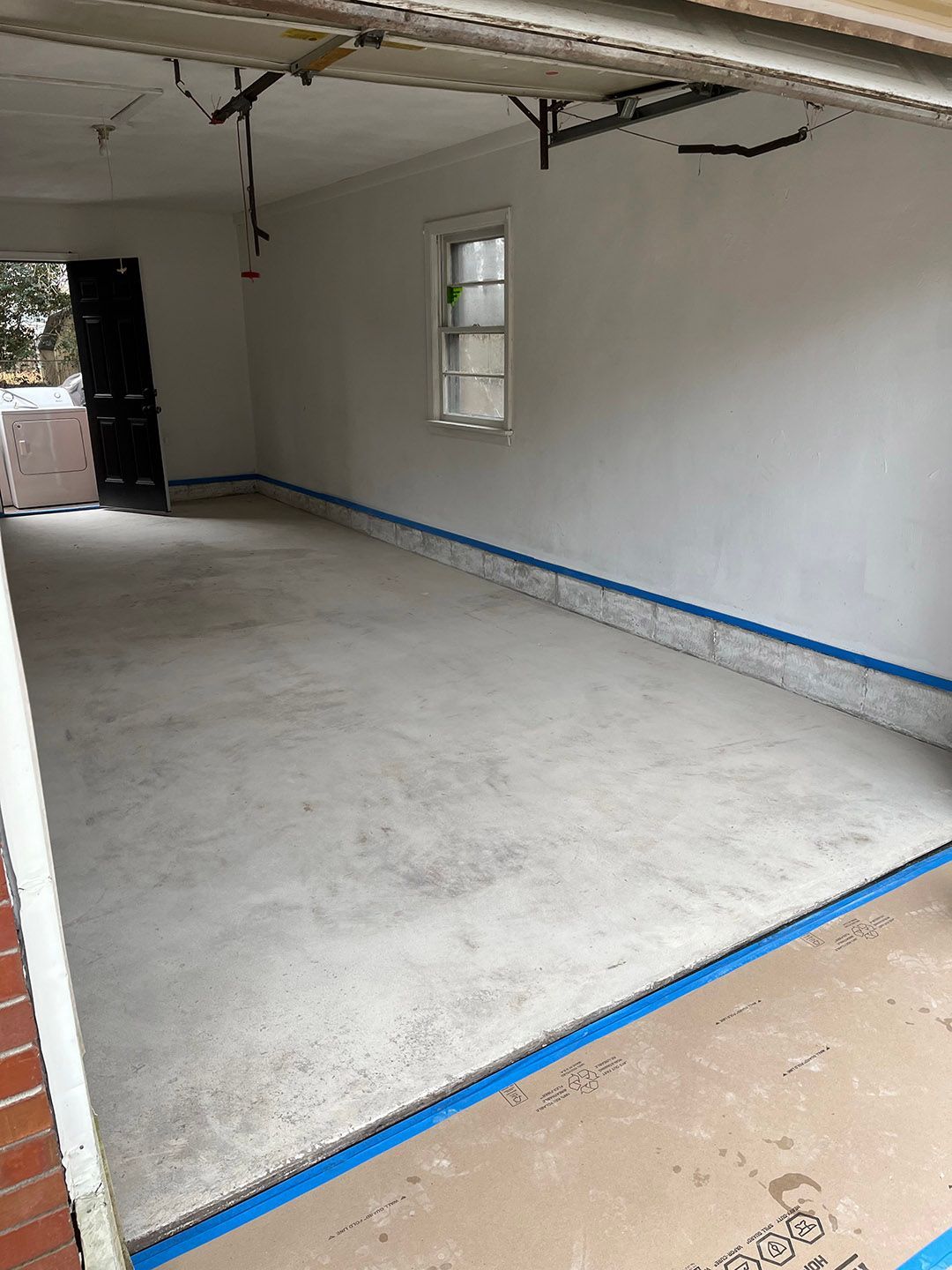 Empty unfinished room with white walls, concrete floor, and blue tape marking the edges.