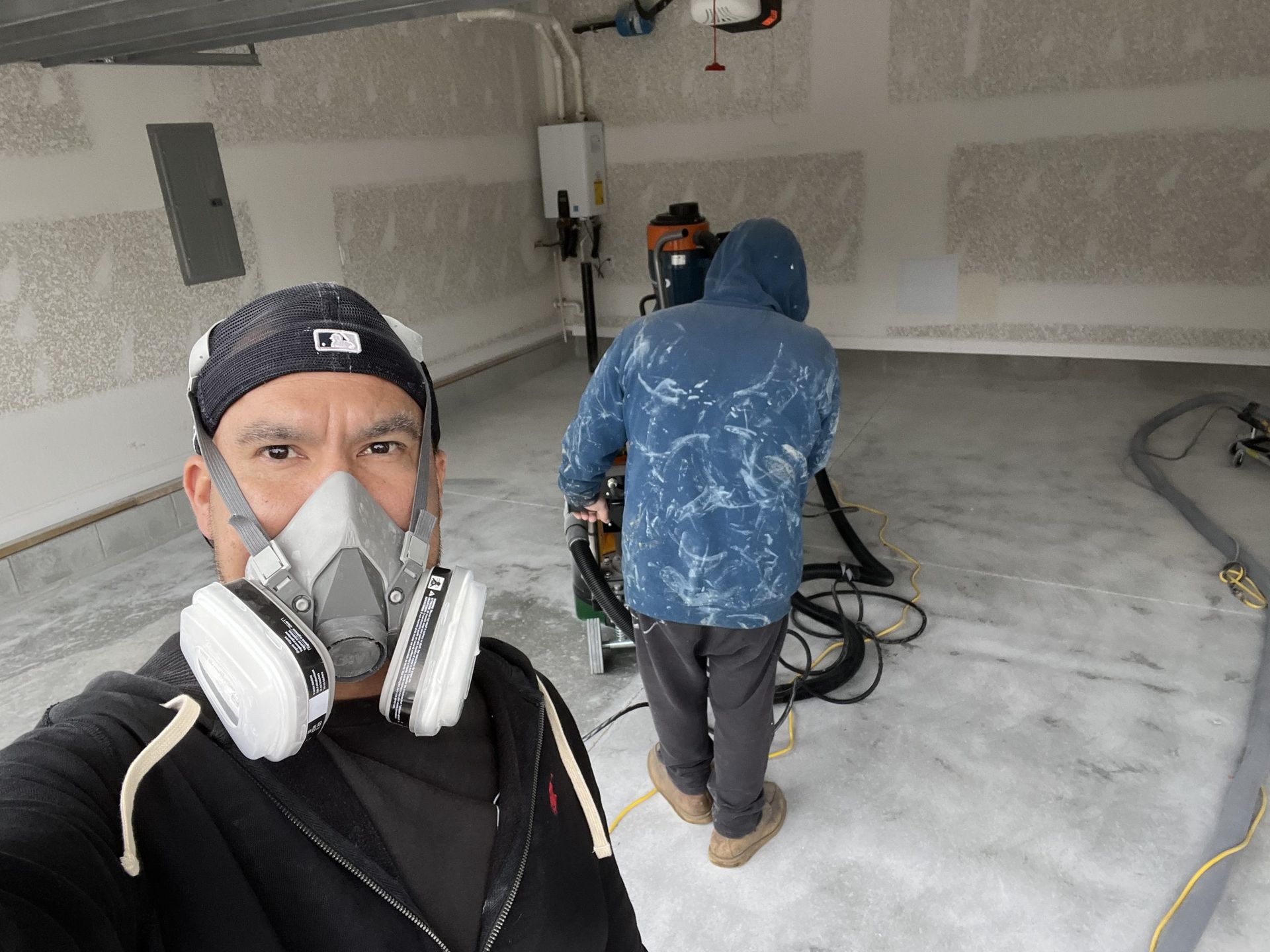 Two workers in a drywall room, one taking a selfie while another operates equipment in the background.