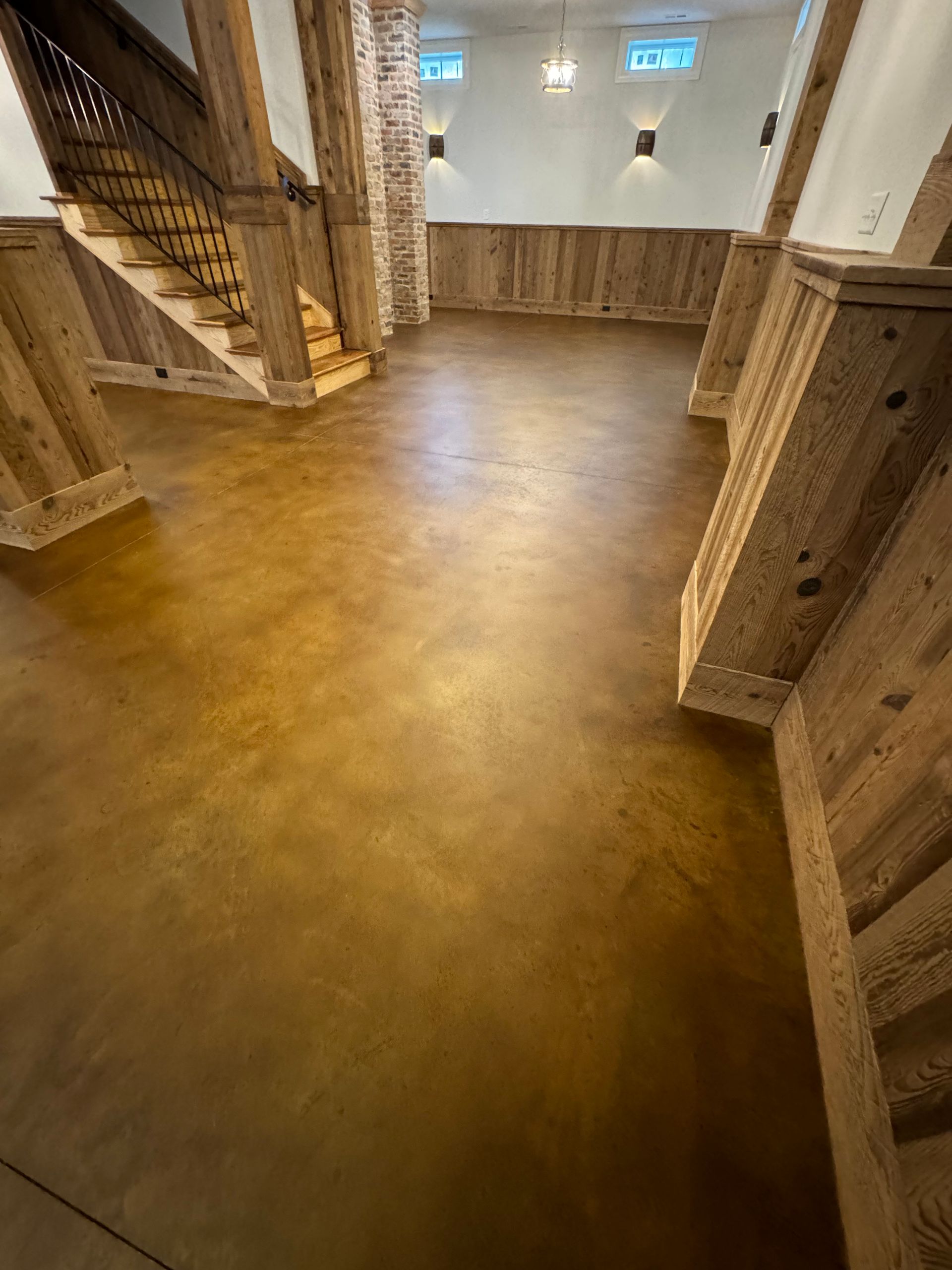 Empty basement room with wood-paneled walls, polished concrete floor, and a staircase on the left.