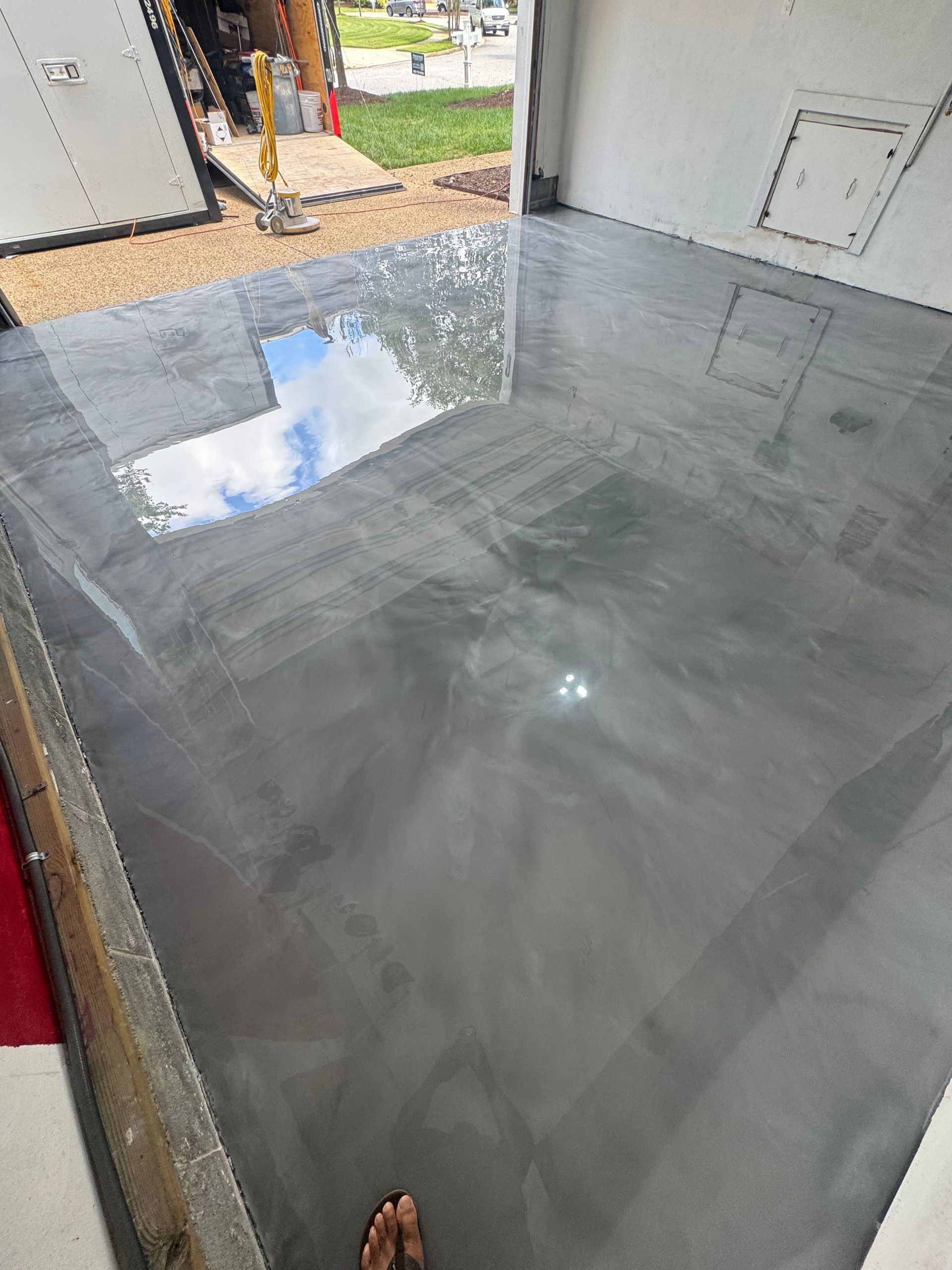 Wet gray concrete floor with reflections inside a garage or shed, with doorway and yard visible outside