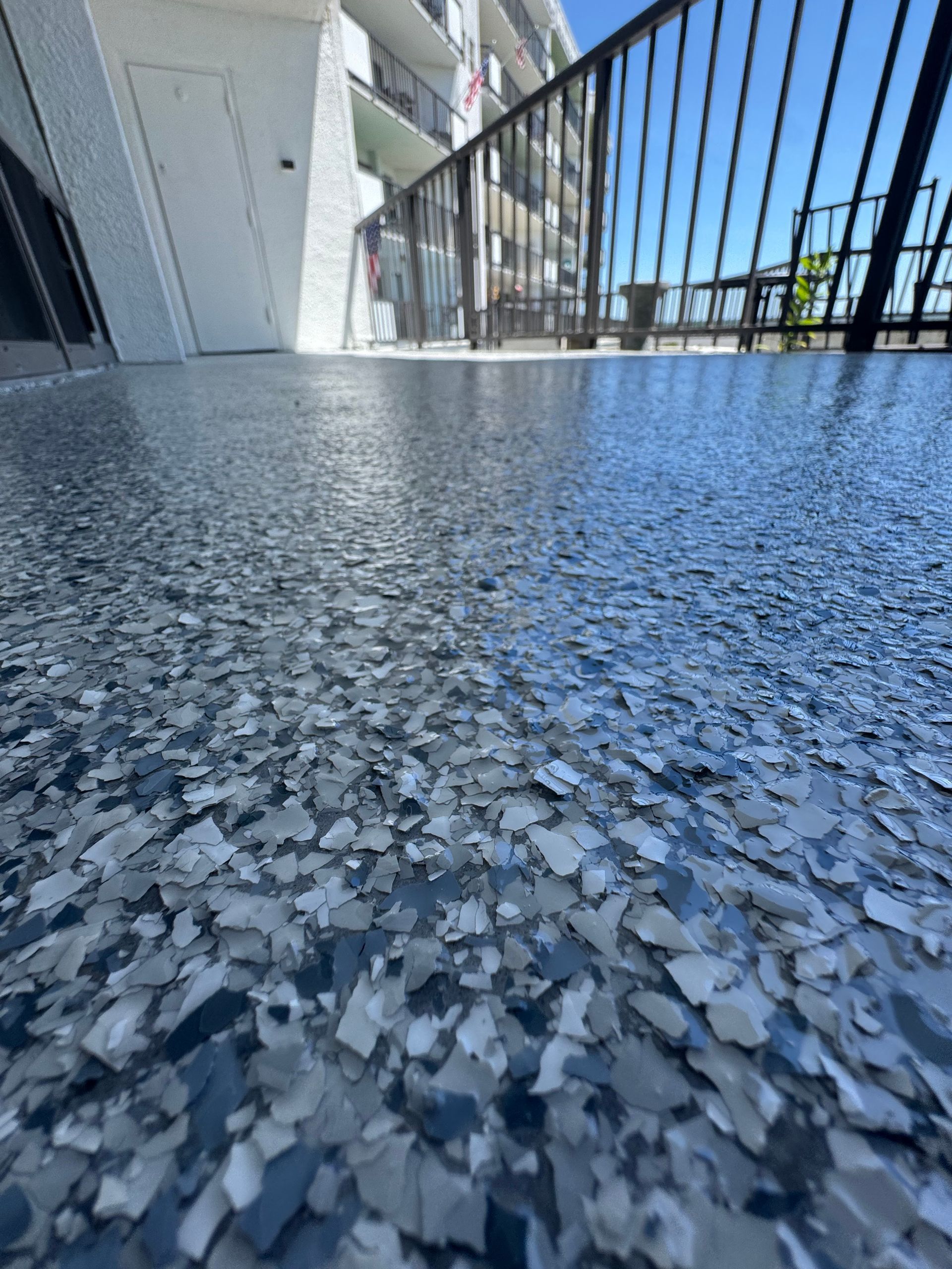 Speckled blue-gray balcony floor with black railing and white wall in bright sunlight
