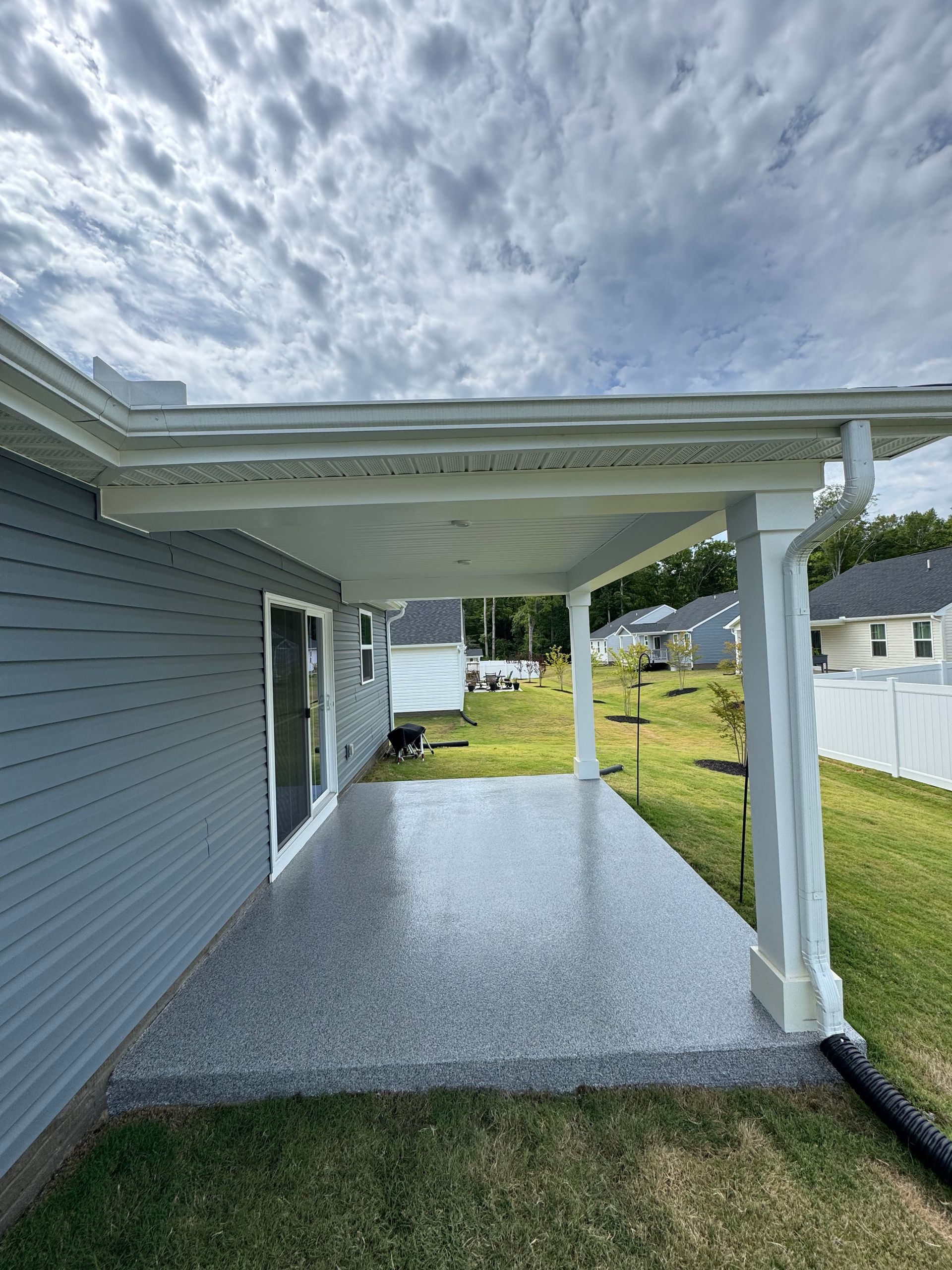 Covered backyard patio with gray siding, white posts, and speckled concrete floor beside a grassy yard.