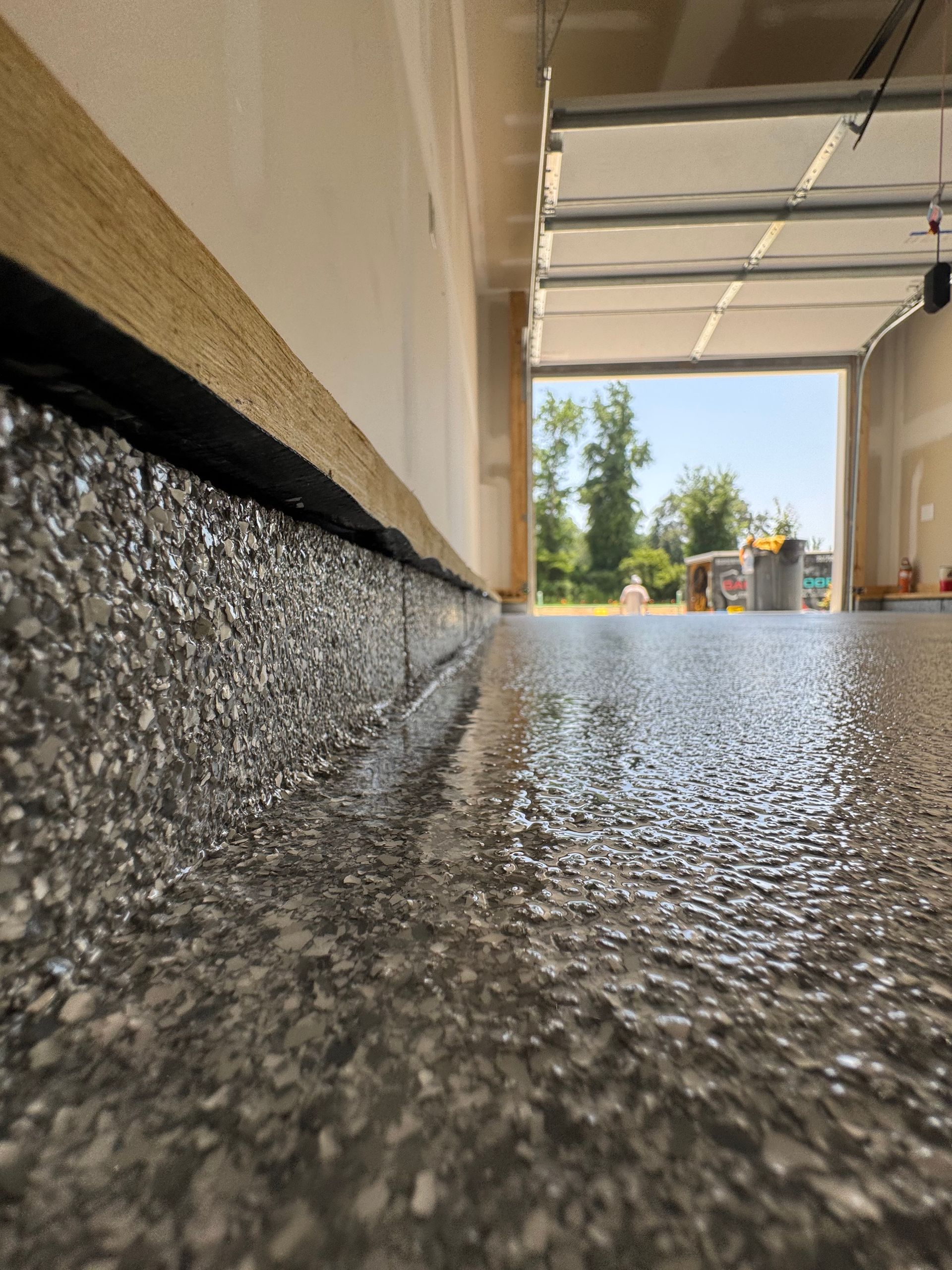 Low-angle view of a wet garage floor leading to an open garage and bright driveway outside