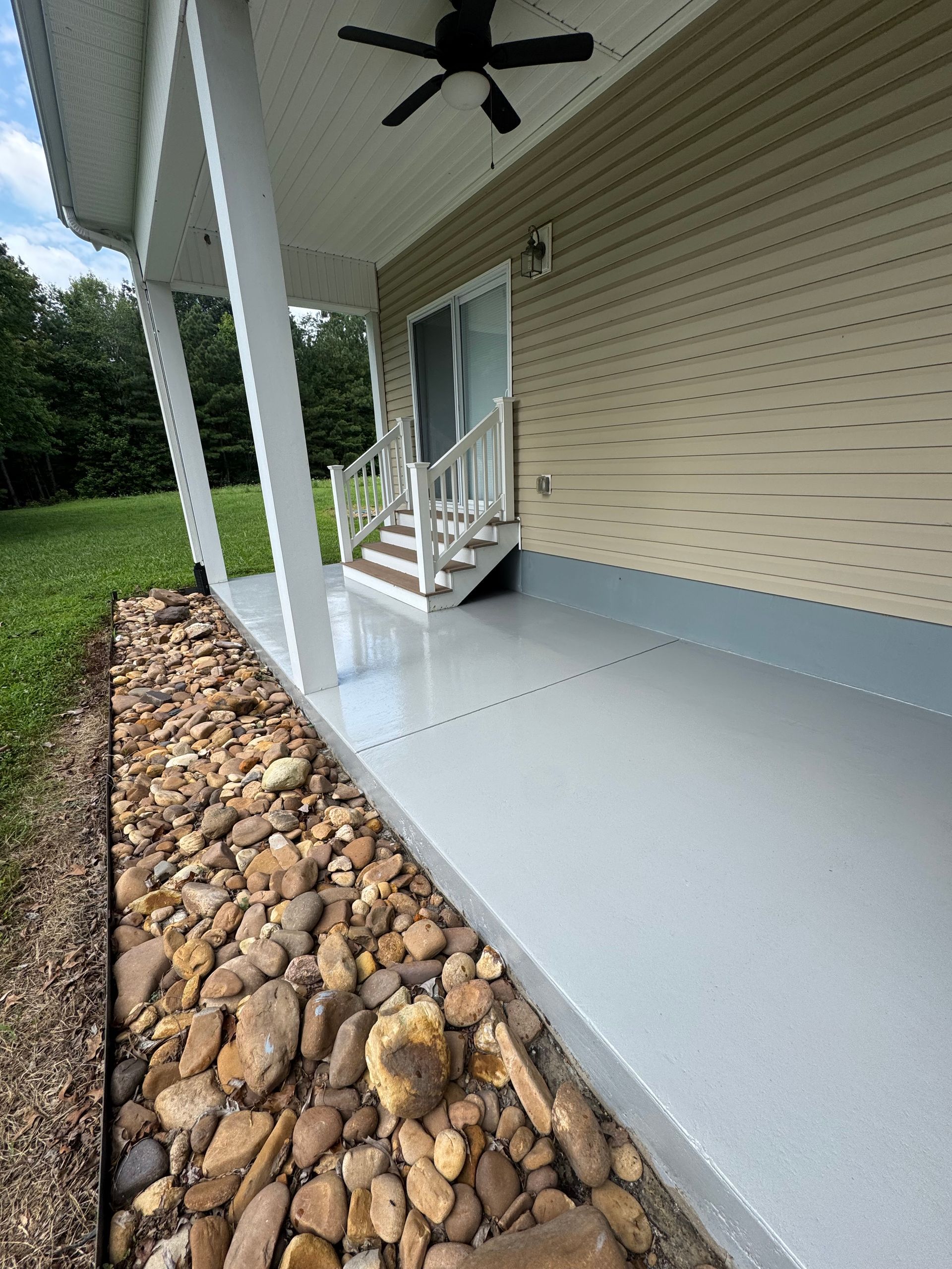 Covered porch with white railing, gray floor, ceiling fan, and stone landscaping along the edge.