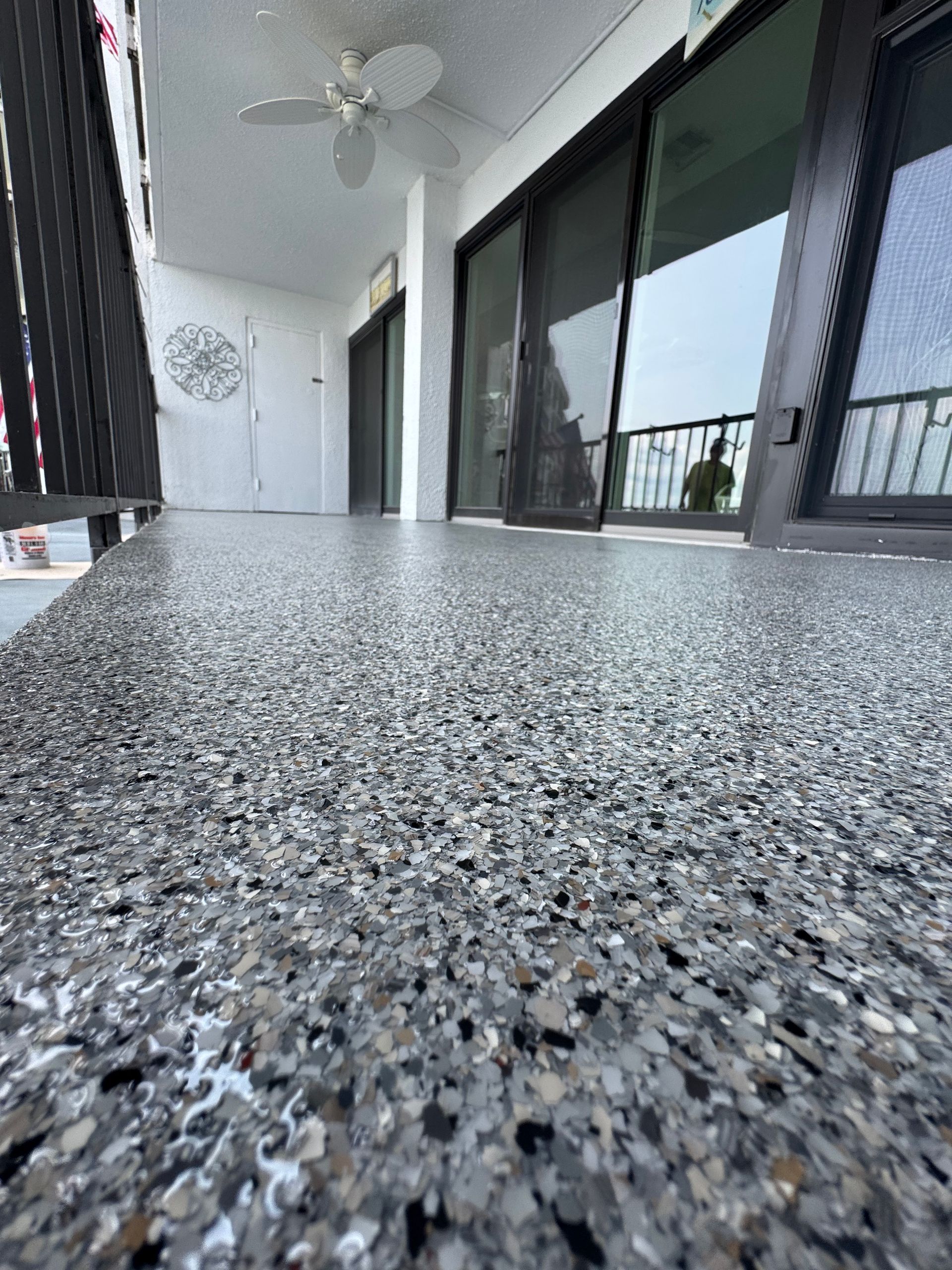 Close-up of a speckled balcony floor with sliding glass doors and a ceiling fan in the background