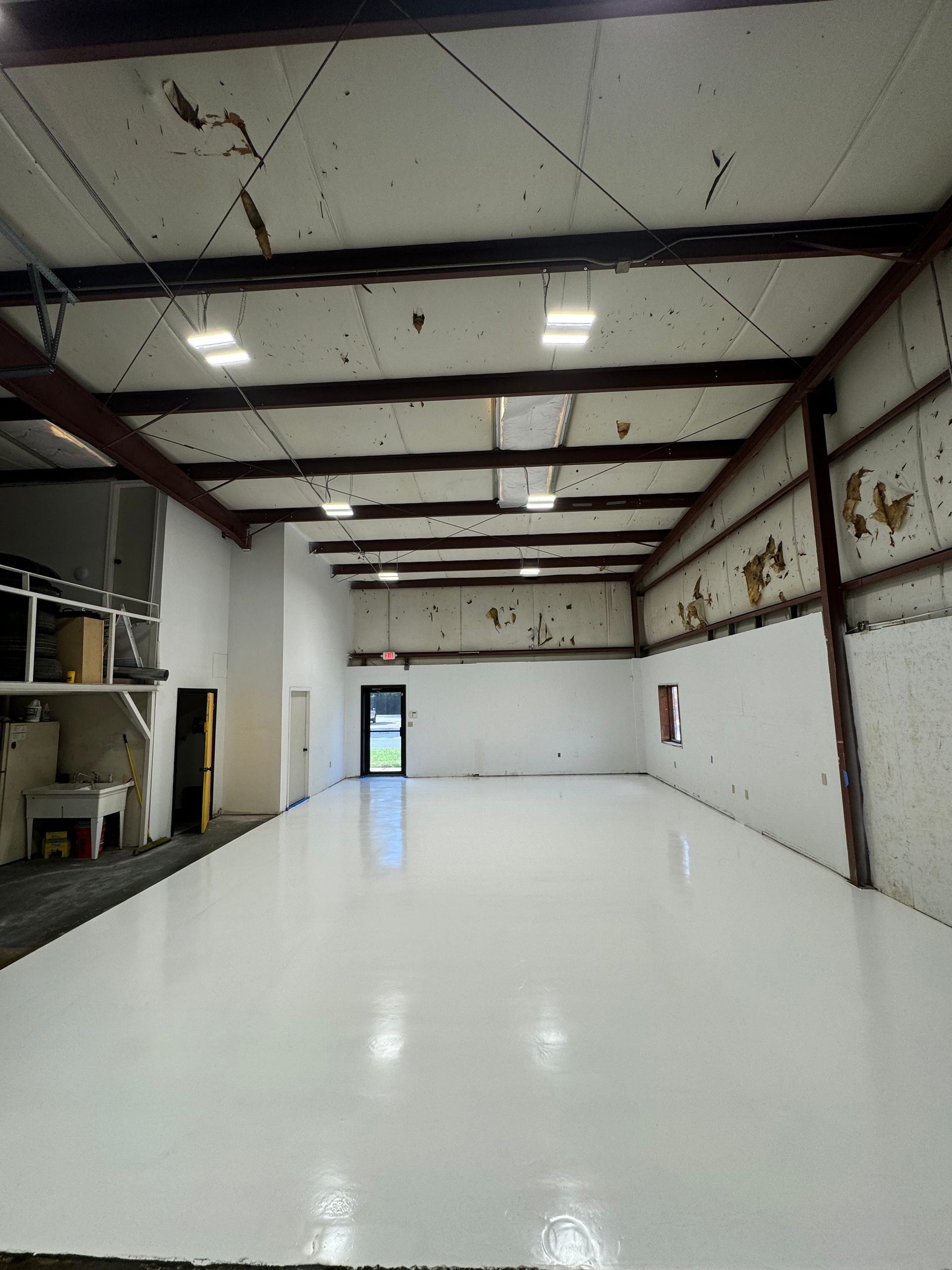 Empty industrial warehouse with white epoxy floor, exposed ceiling beams, and open doorway at the far end