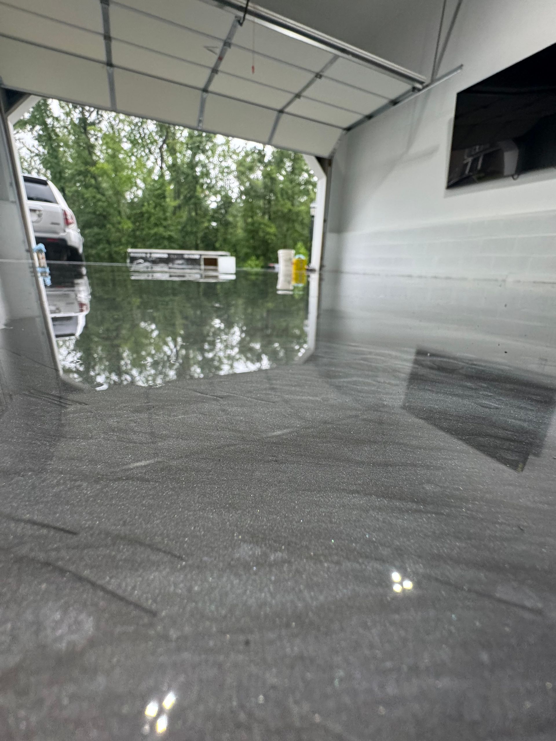 Flooded garage interior with standing water reflecting the open door and parked vehicles outside