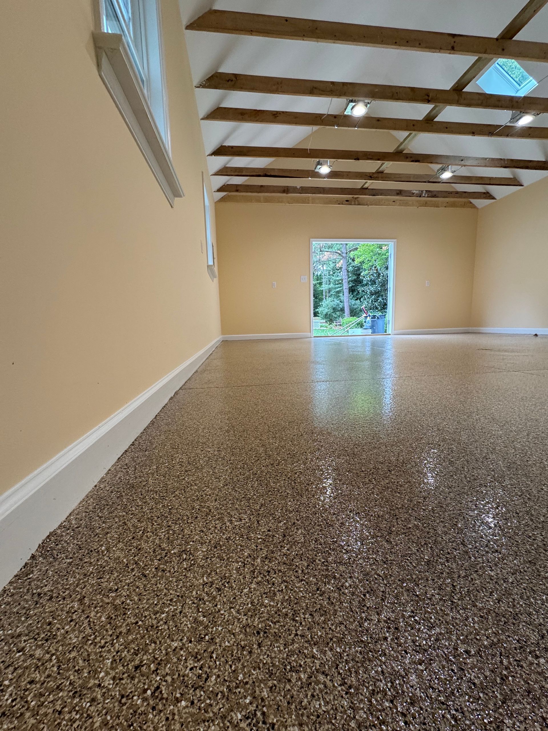 Empty beige room with exposed wood beams, speckled floor, and open doorway to greenery