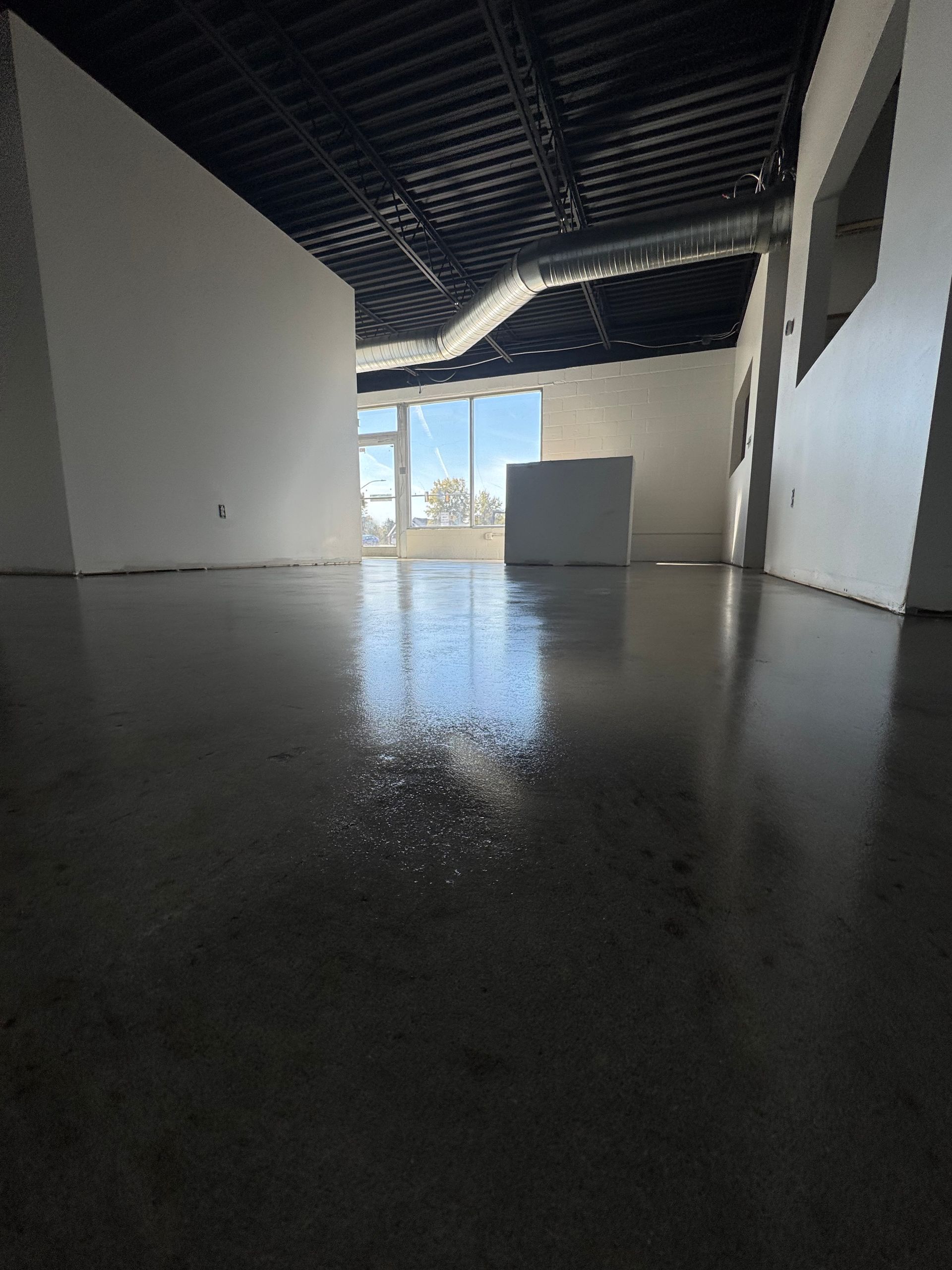 Empty unfinished room with glossy concrete floor, white walls, exposed ceiling, and bright windows at the far end