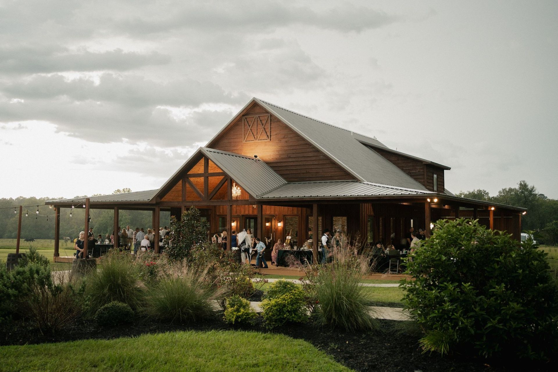 Rustic house with a covered porch and people gathered outside in a green countryside setting at dusk