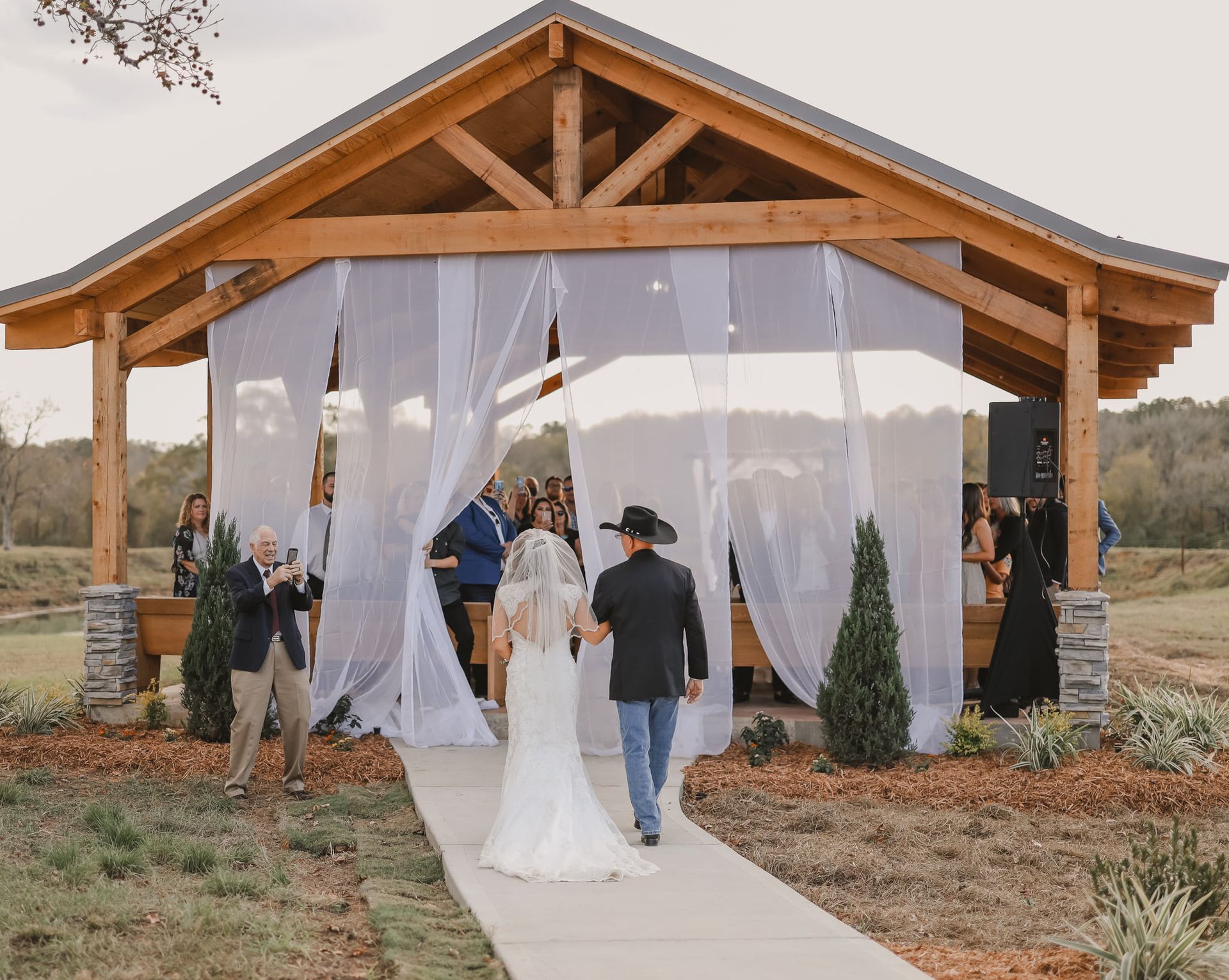 Couple walks up a path to a rustic wedding pavilion with sheer white curtains and guests nearby