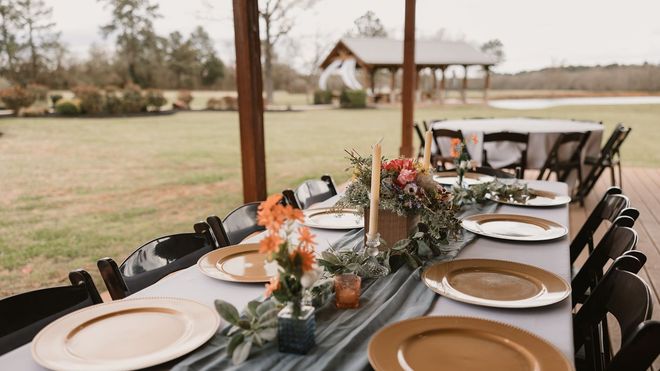 Outdoor rustic dining table set with white and gold plates, floral centerpieces, under a covered patio overlooking a lawn