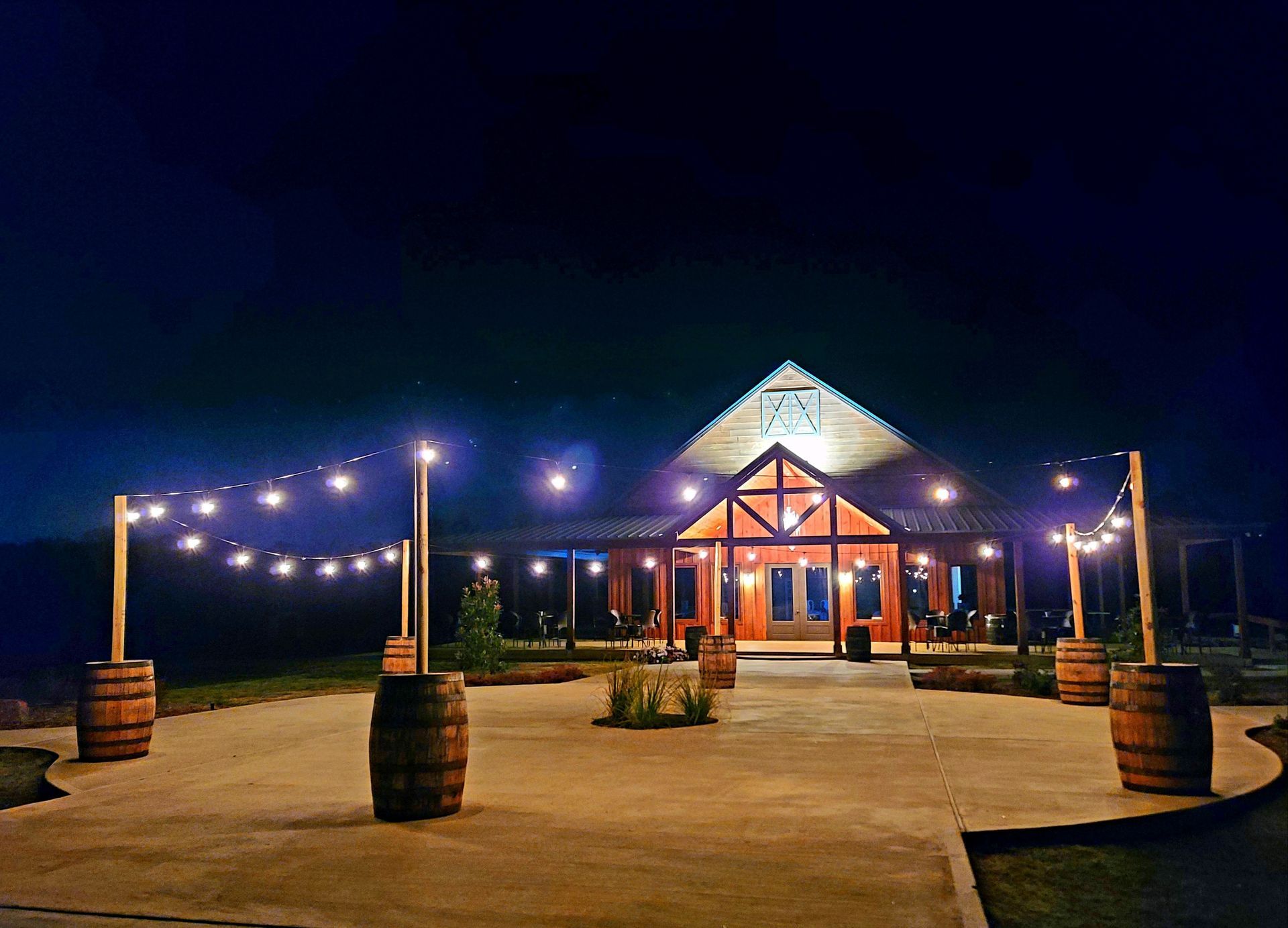 Nighttime view of a lit pavilion with string lights and barrel planters in a paved outdoor courtyard