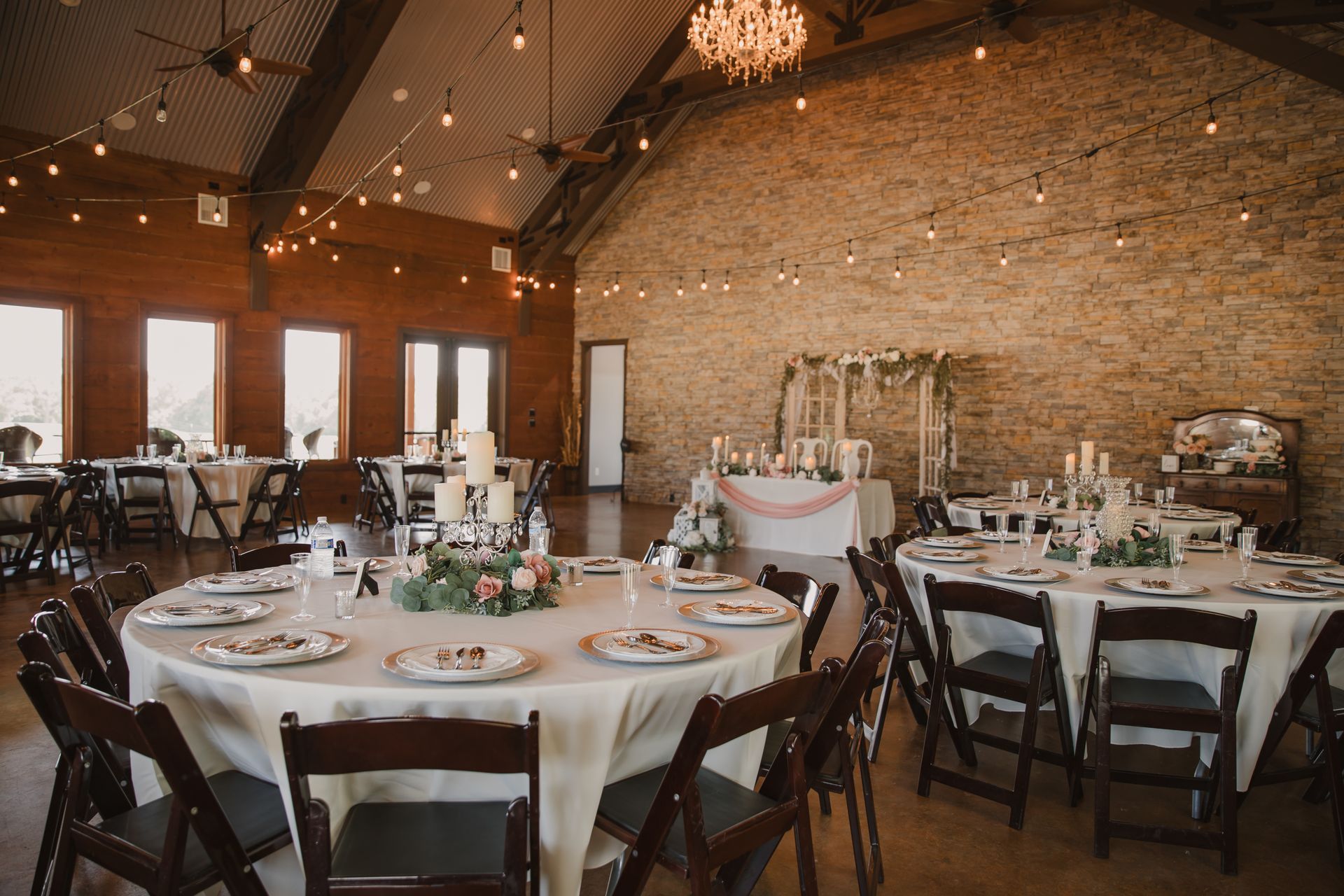 Rustic banquet hall with round, set dining tables and chairs under warm string lights and exposed brick walls