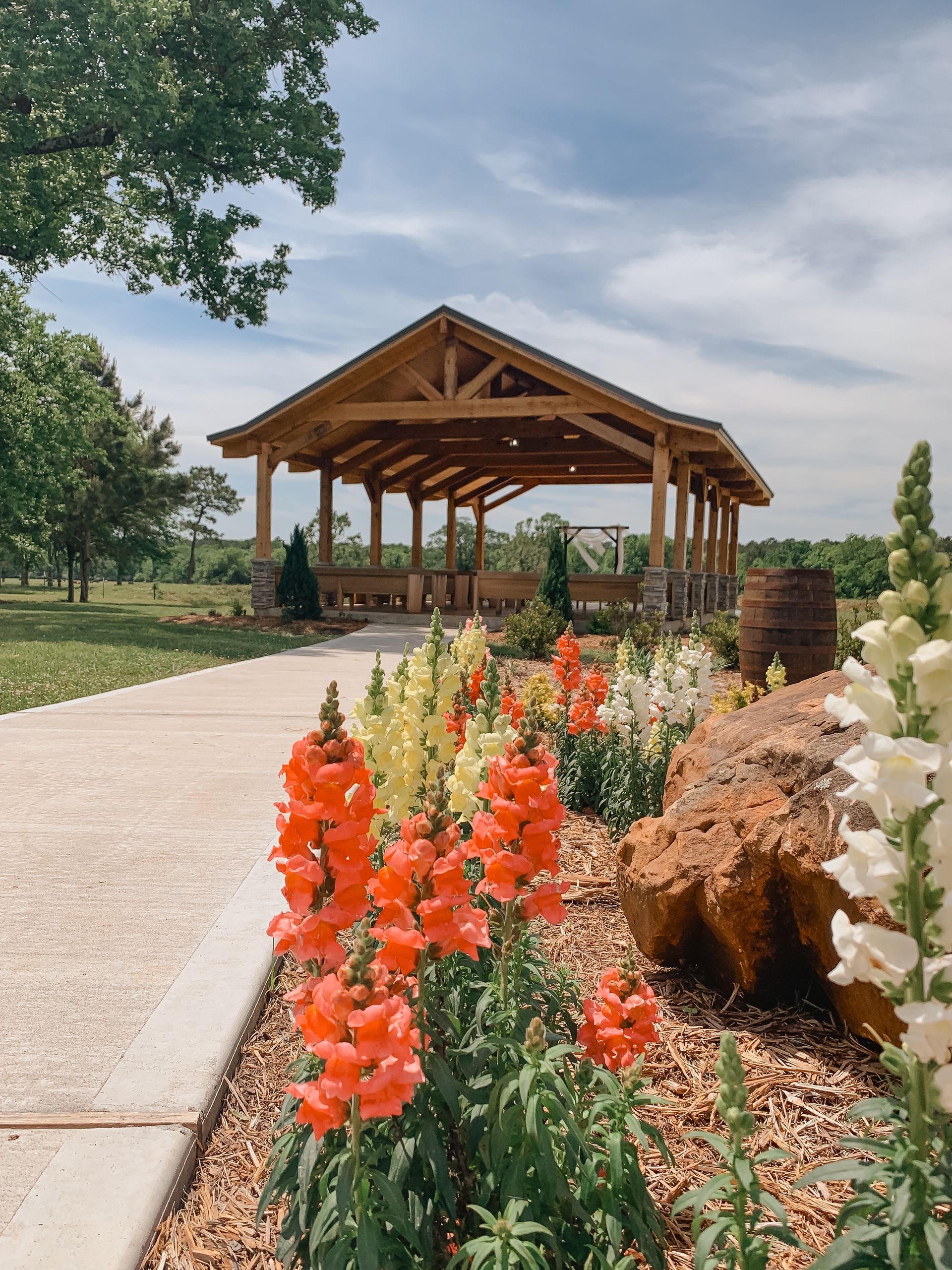 Wooden pavilion beside a paved path with red flowers and landscaped greenery under a cloudy sky