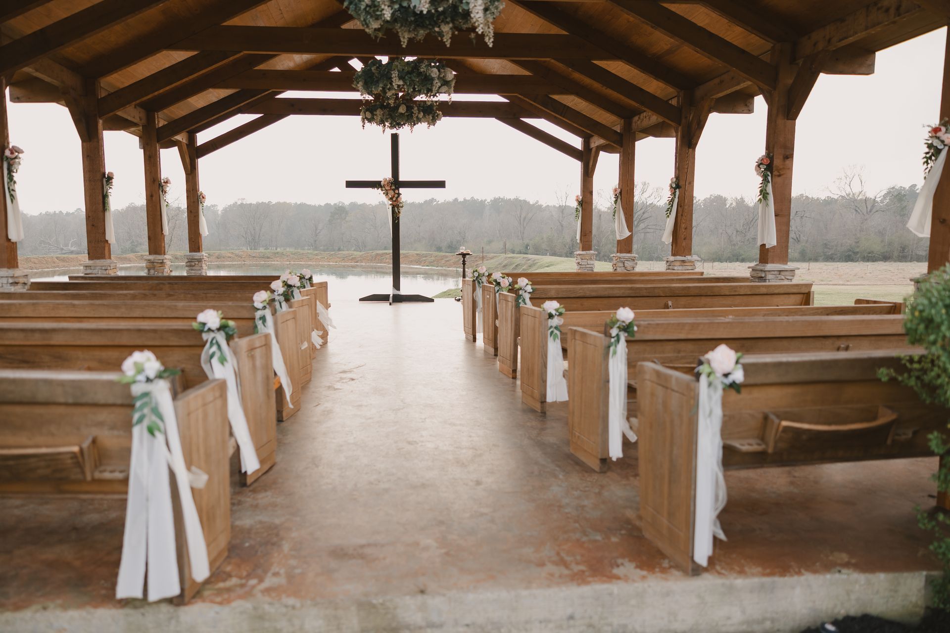 Empty rustic wedding pavilion with decorated benches and central aisle facing a cross at the altar