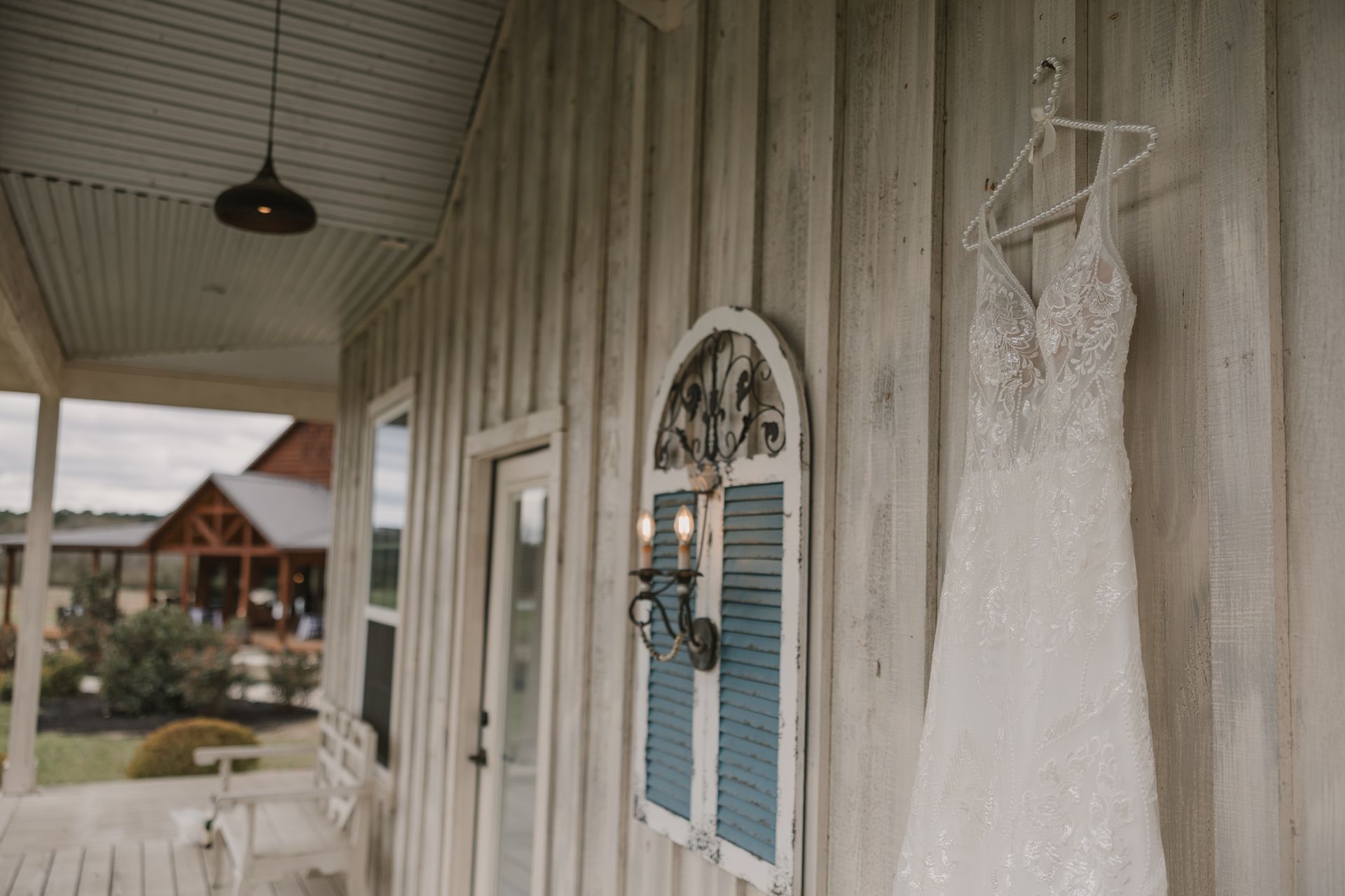 White lace wedding dress hanging on a wooden porch wall beside a blue shuttered window and mirror.