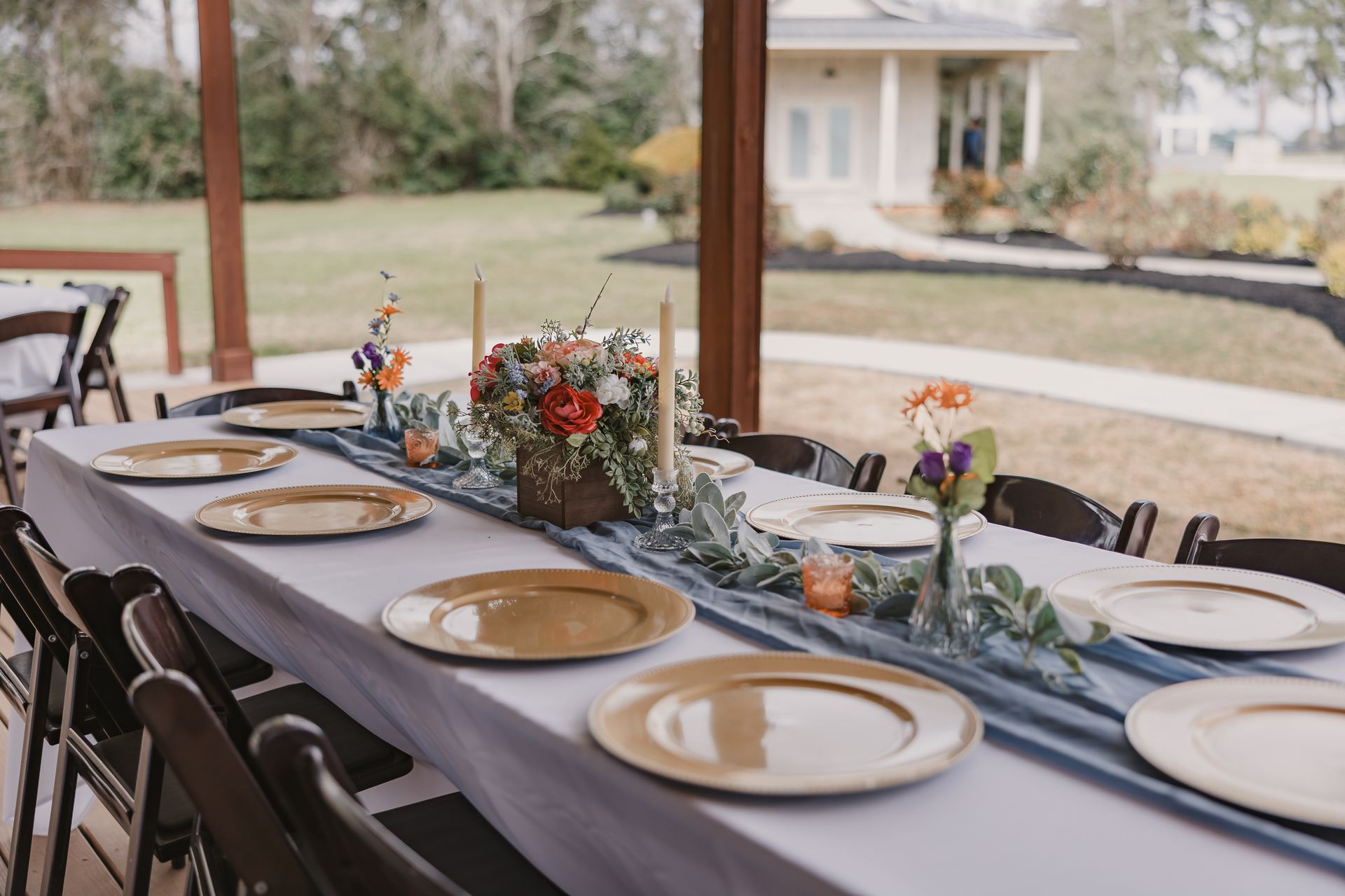 Decorated outdoor dining table with white runner, gold plates, and floral centerpieces under a patio
