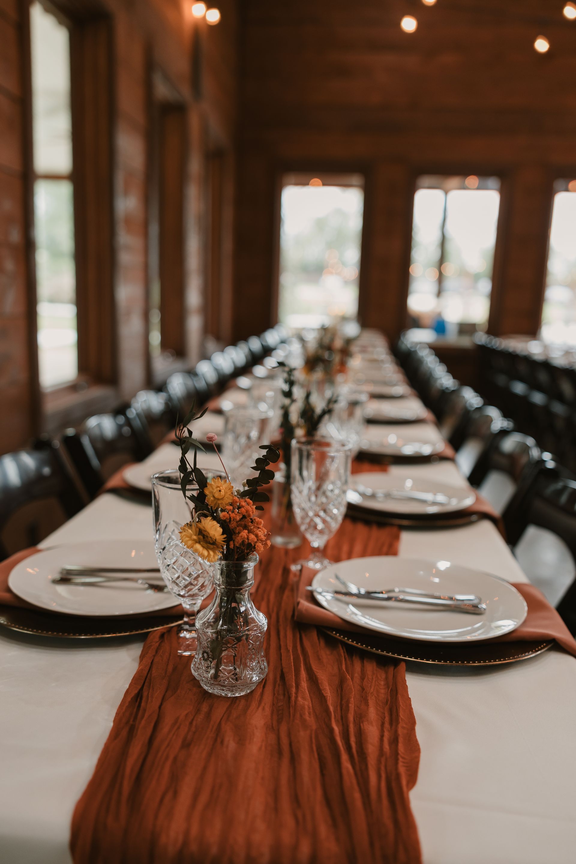 Long banquet table set with white plates, glassware, and orange floral centerpieces in a wooden hall