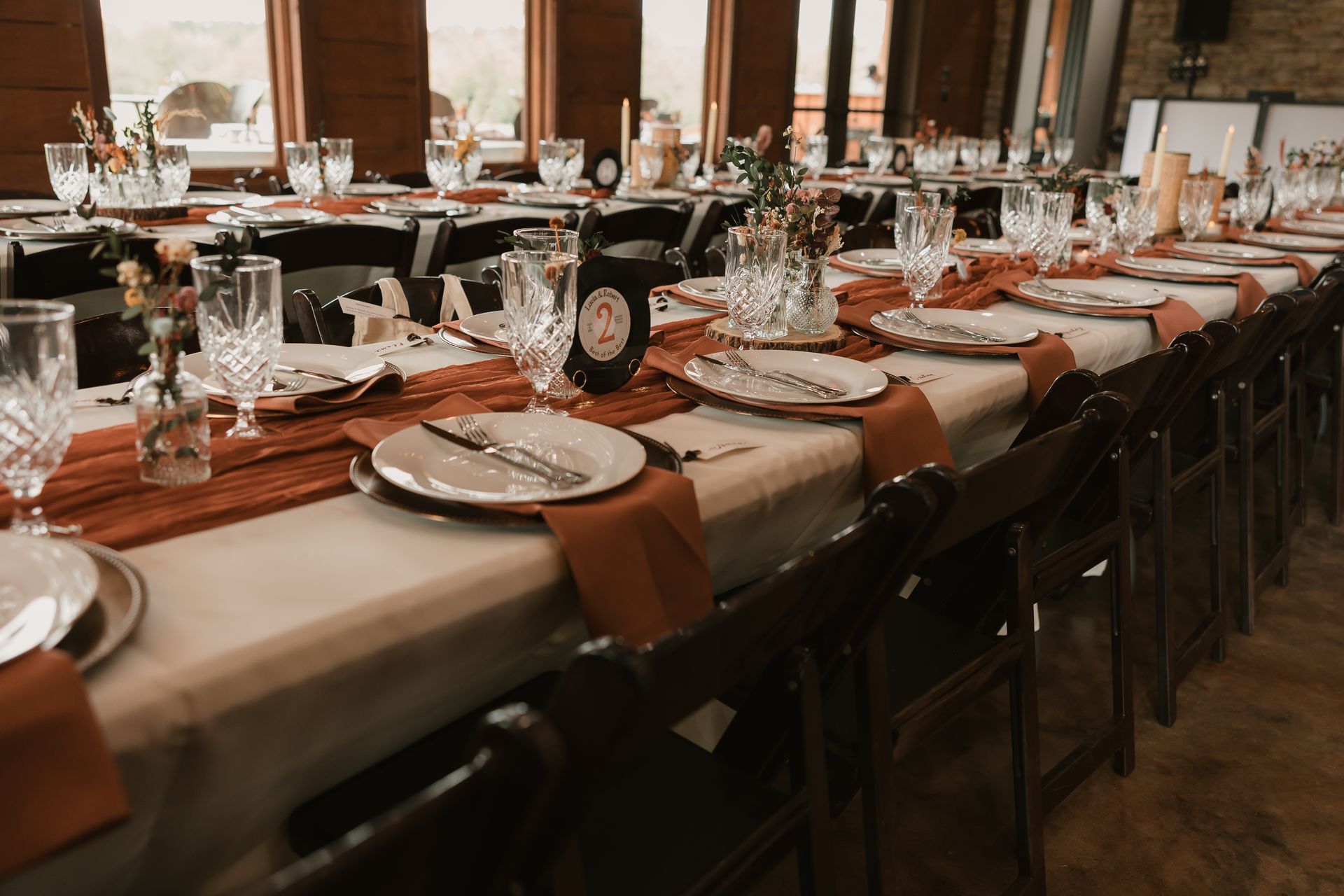Long banquet table set with white plates, glassware, and burnt orange napkins in a warm-lit dining room