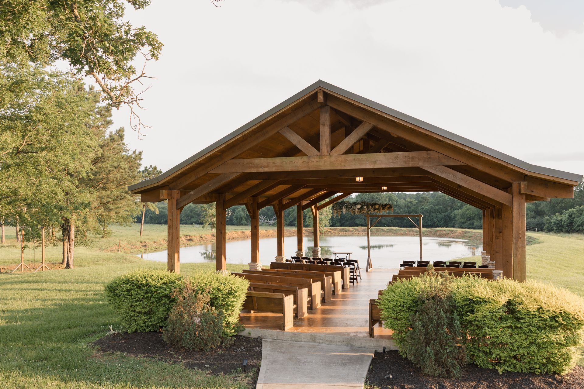 Wooden lakeside pavilion with picnic tables, framed by trimmed hedges and trees