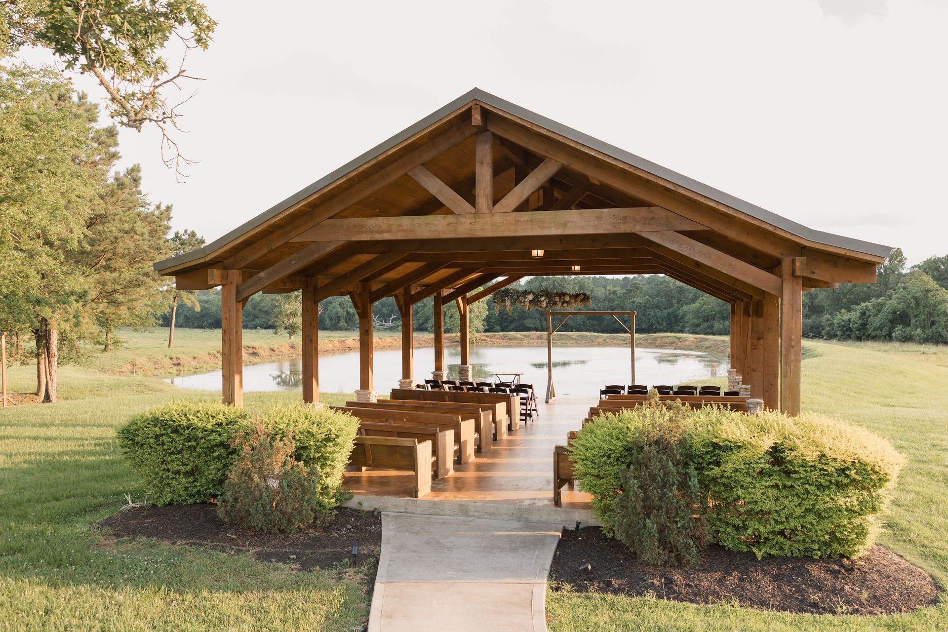 Wooden lakeside pavilion with picnic tables and benches, surrounded by grass and bushes