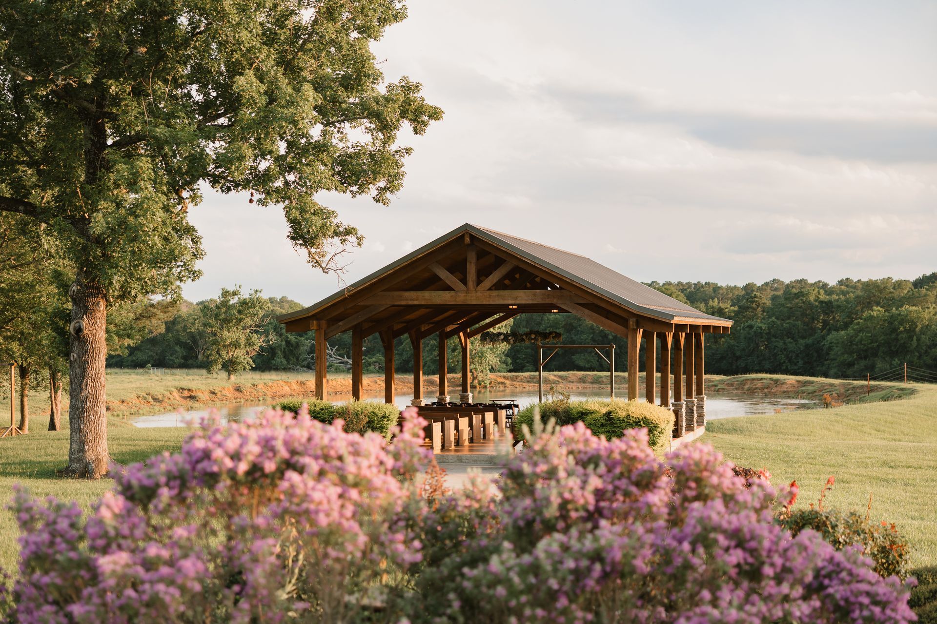 Wooden pavilion by a lake, framed by pink flowers and trees at sunset