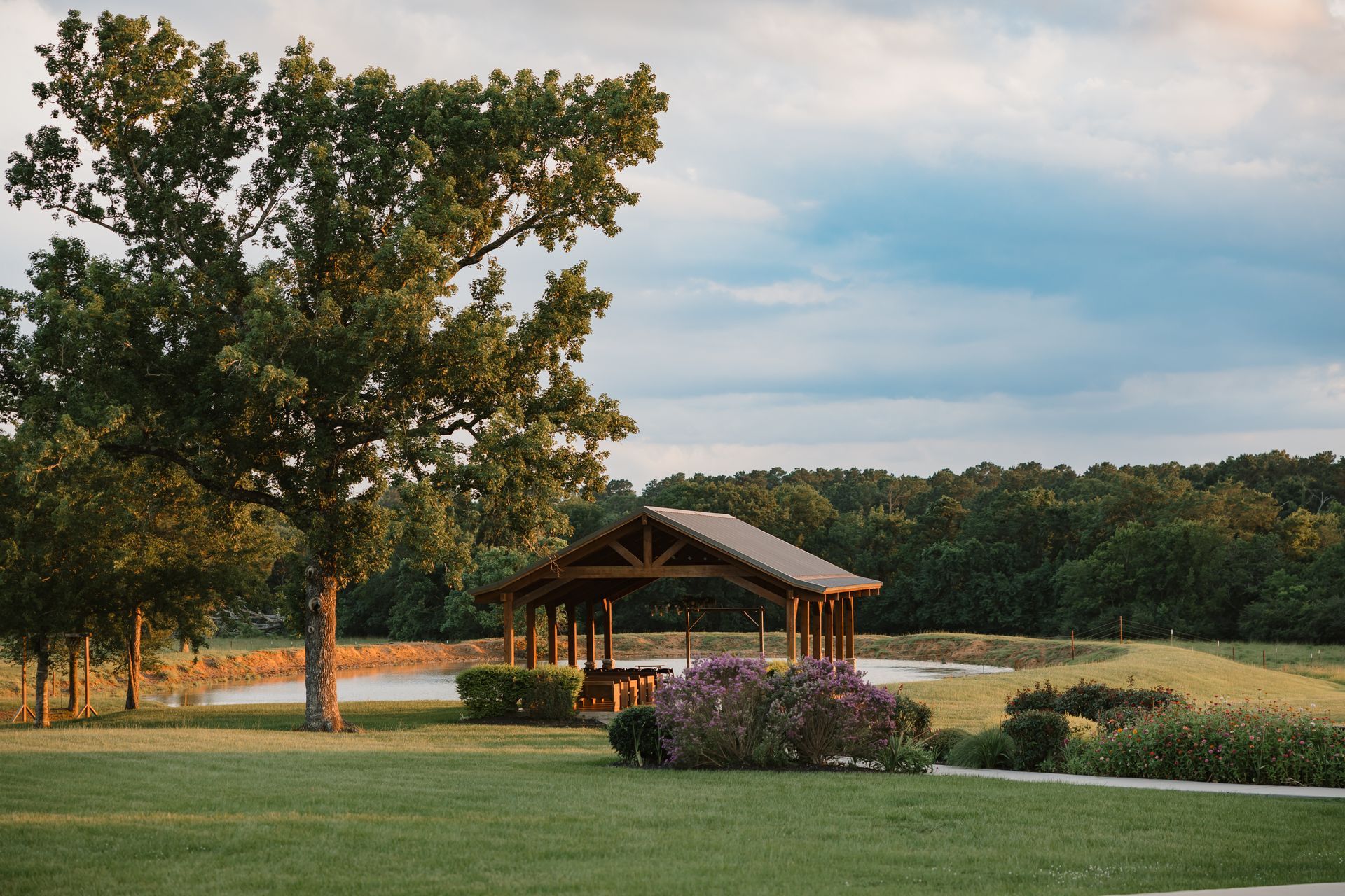 Gazebo by a pond in a grassy field at sunset, with trees and purple flowers in the foreground