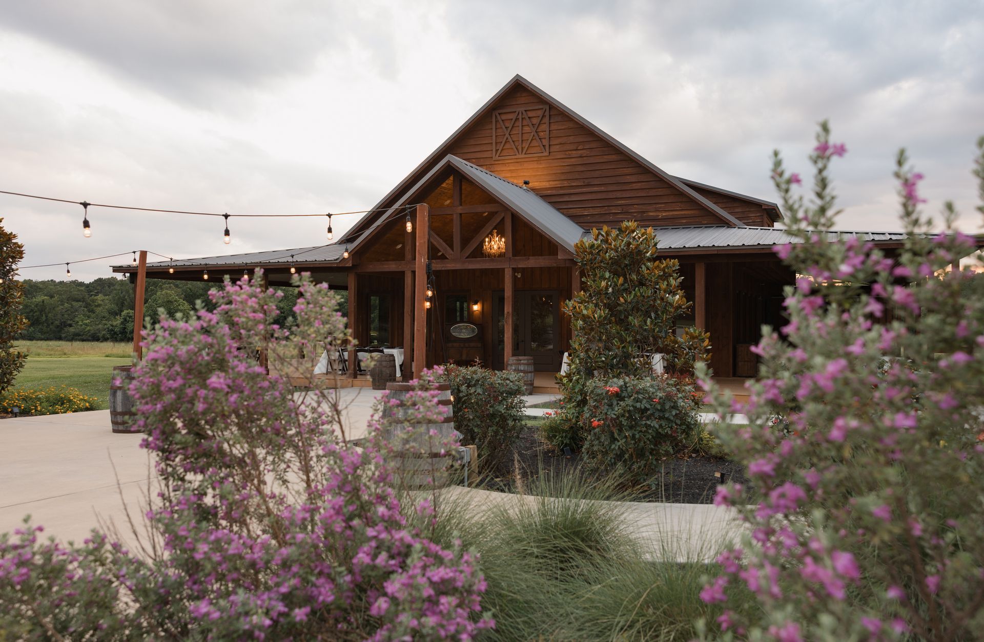 Rustic wooden cabin with a peaked roof, surrounded by pink flowers and greenery under a clear sky