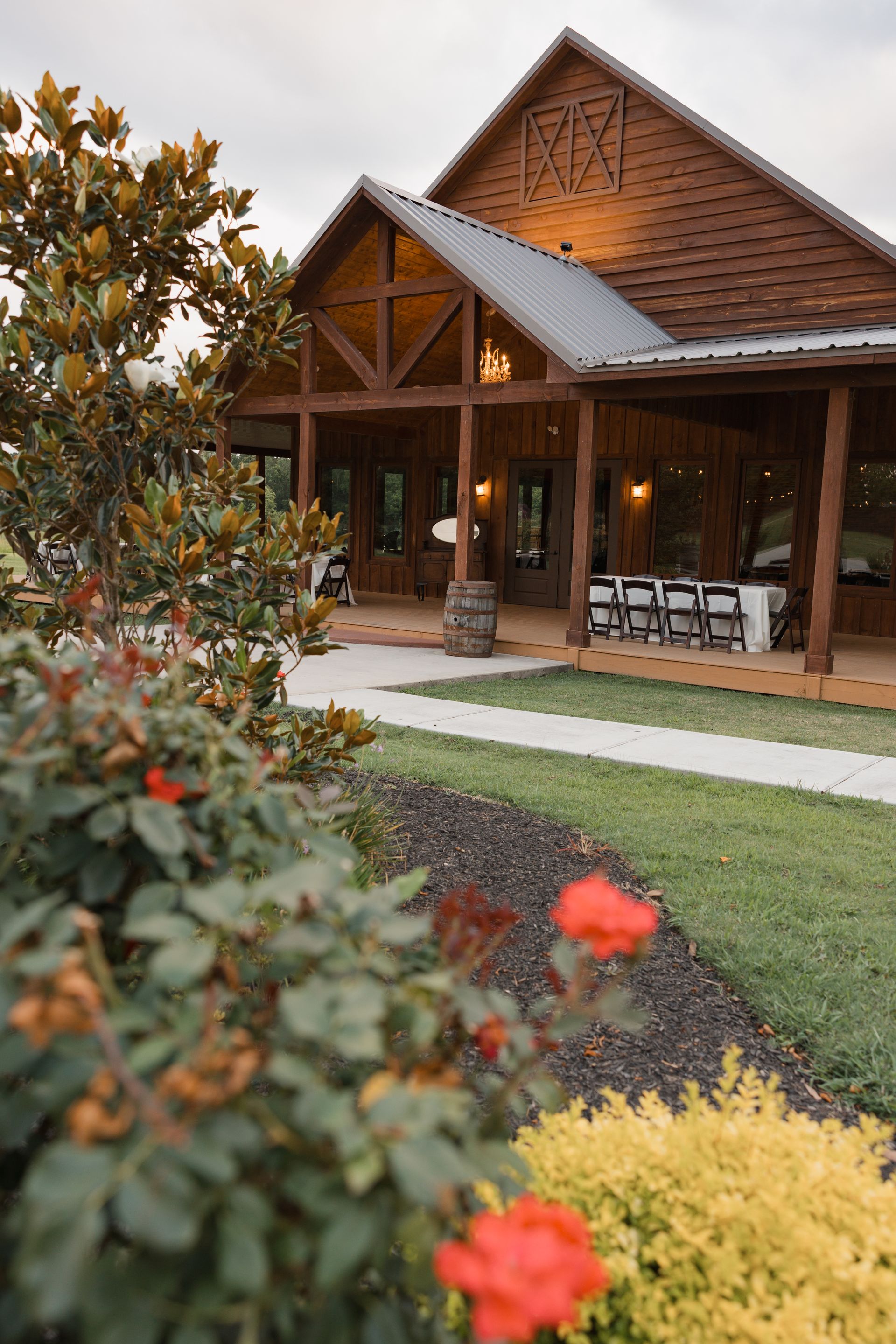 Rustic wooden house with porch and patio furniture, framed by red flowers and a green lawn