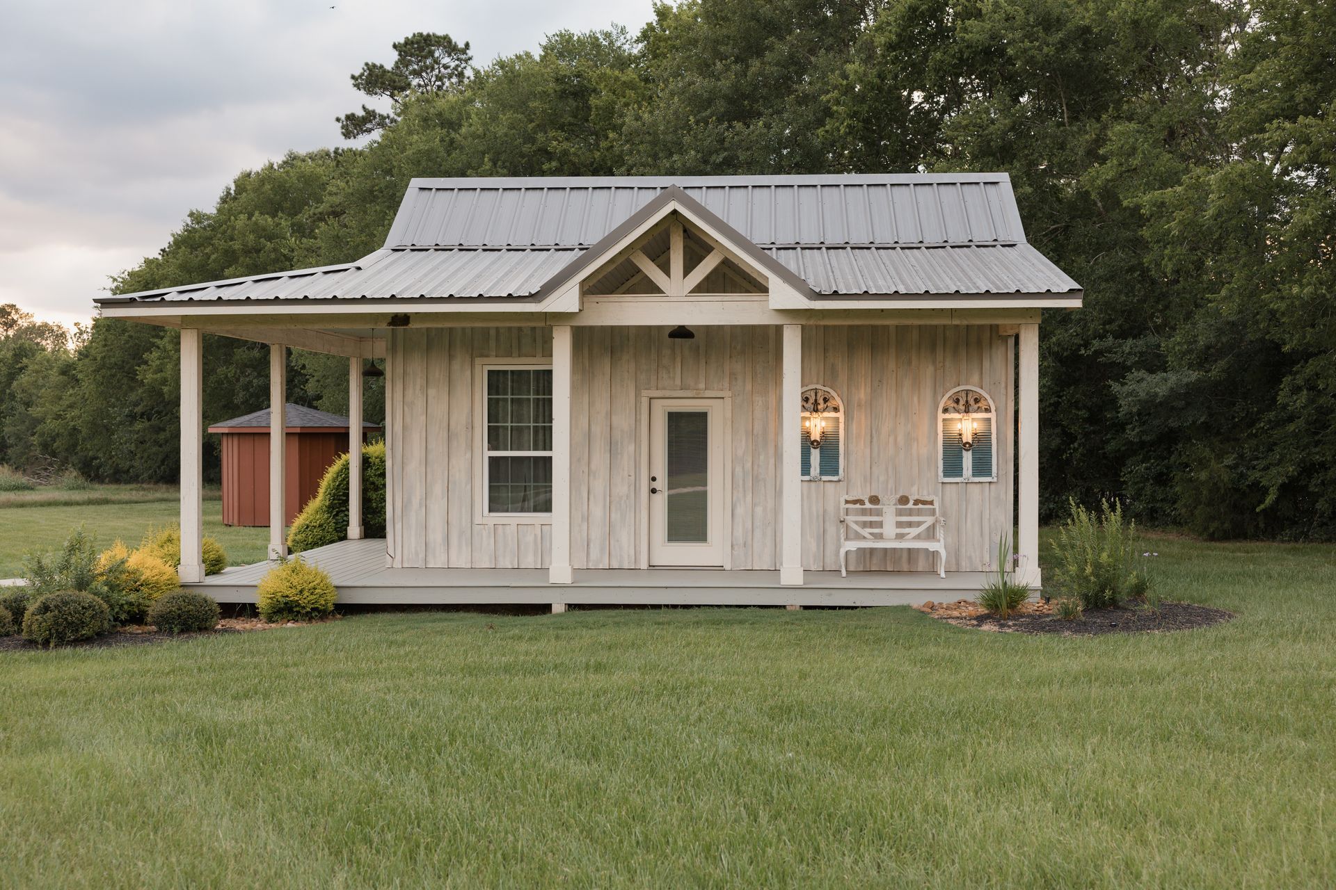 Small light-gray cottage with metal roof and porch in a grassy yard at dusk