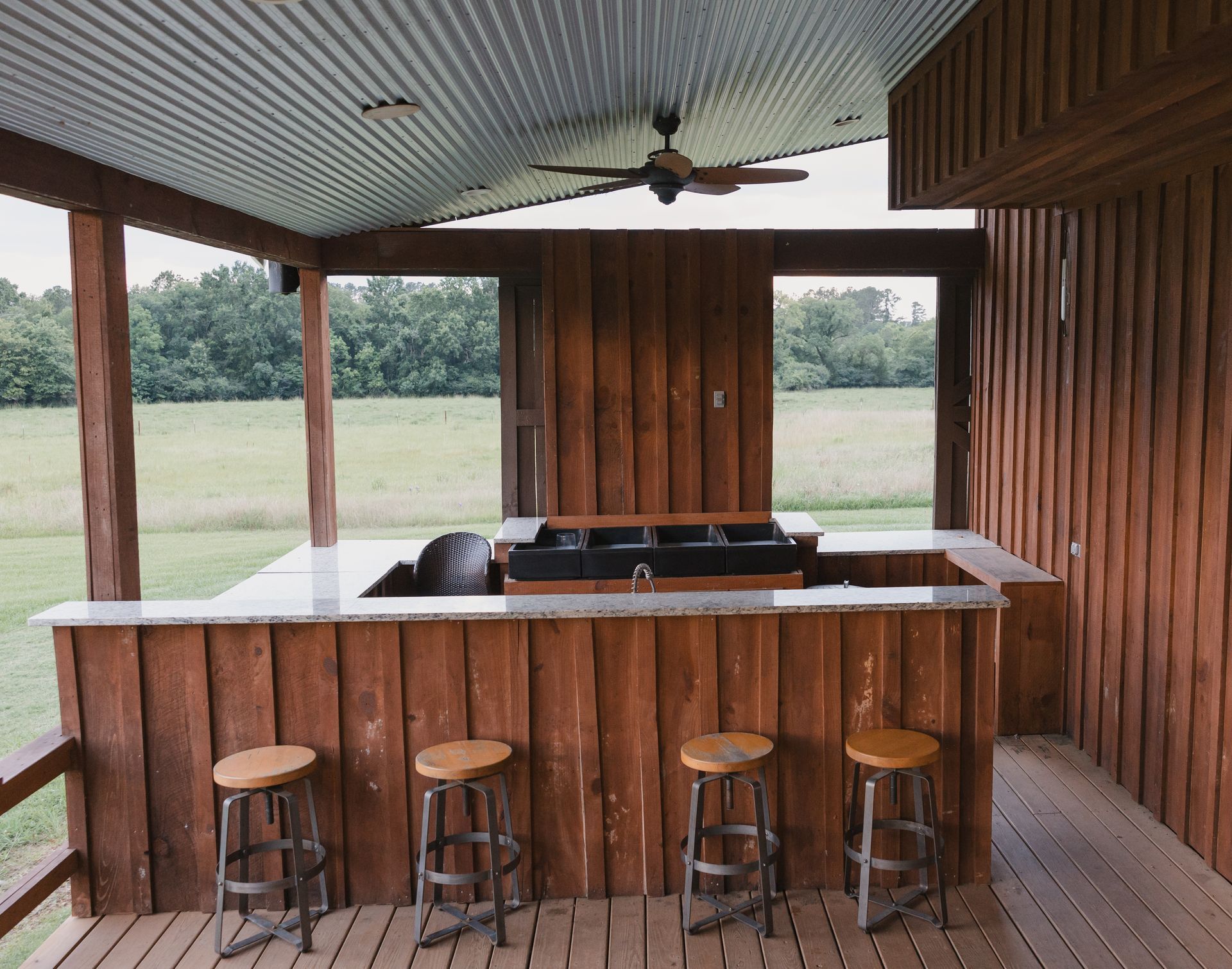 Rustic outdoor bar with stools under a covered wooden pavilion overlooking a grassy field