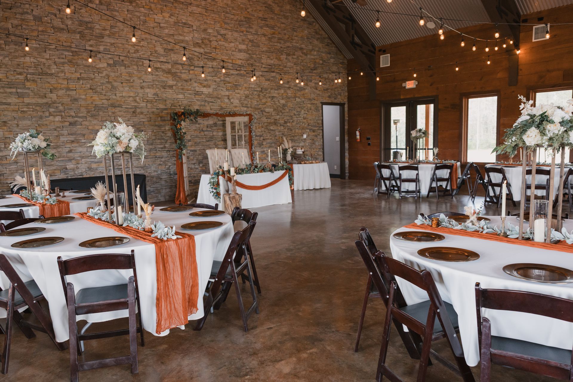 Rustic banquet hall set with round tables, white tablecloths, orange runners, and dark wooden chairs under string lights
