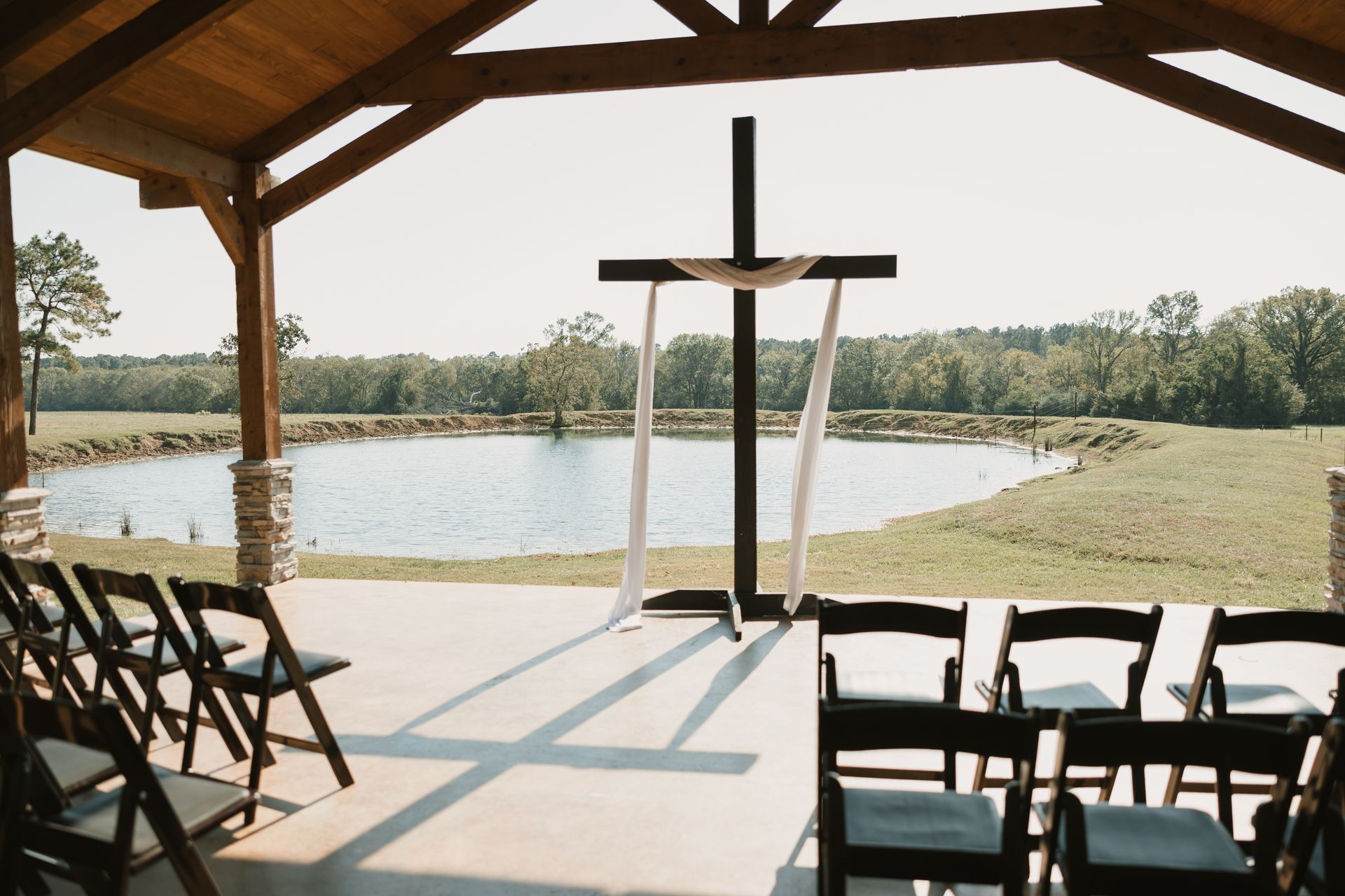Outdoor wedding ceremony area with cross, rows of chairs, and a lake view under a wooden pavilion