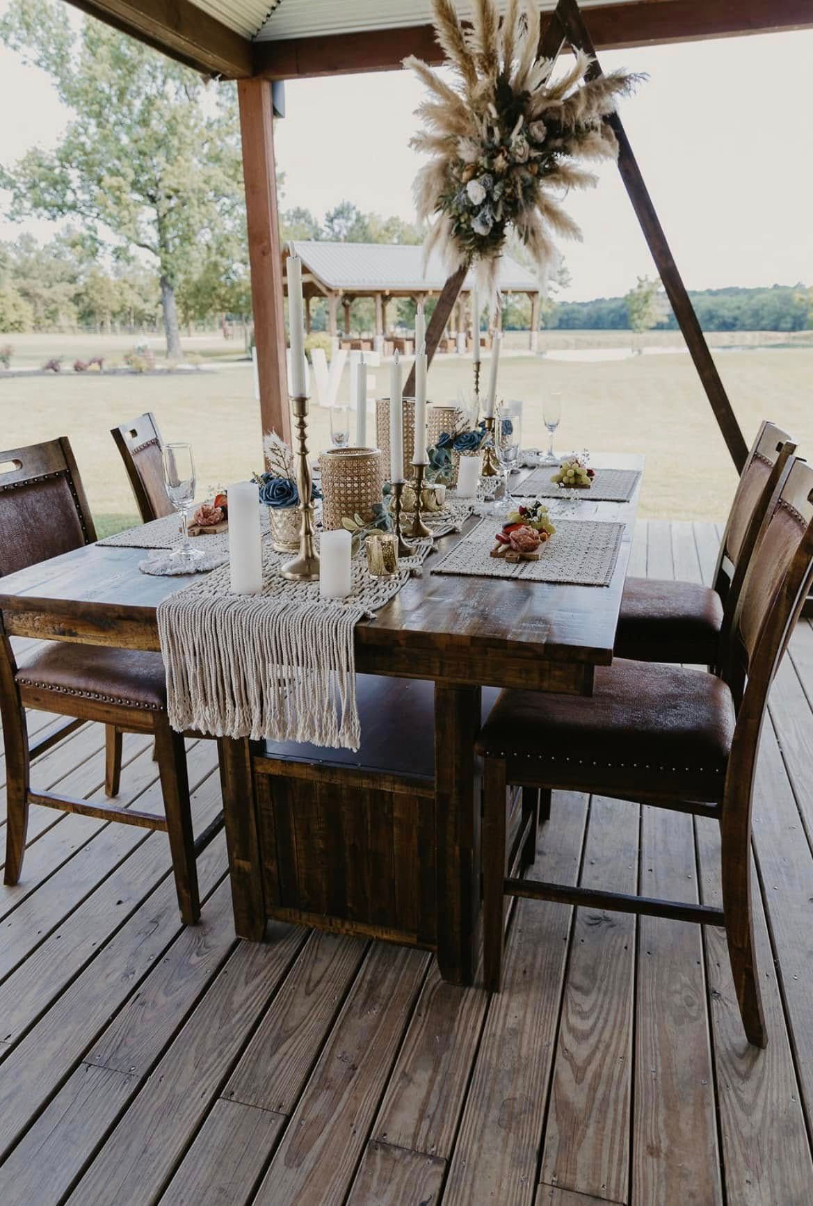 Outdoor dining table on a wooden deck, set with candles, flowers, and place settings by the water.