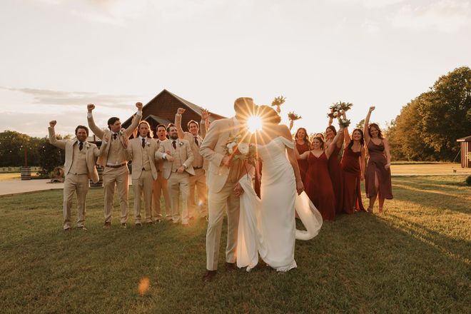 Wedding party walking on a grassy lawn at sunset, with a barn in the background and sunlight flaring behind them
