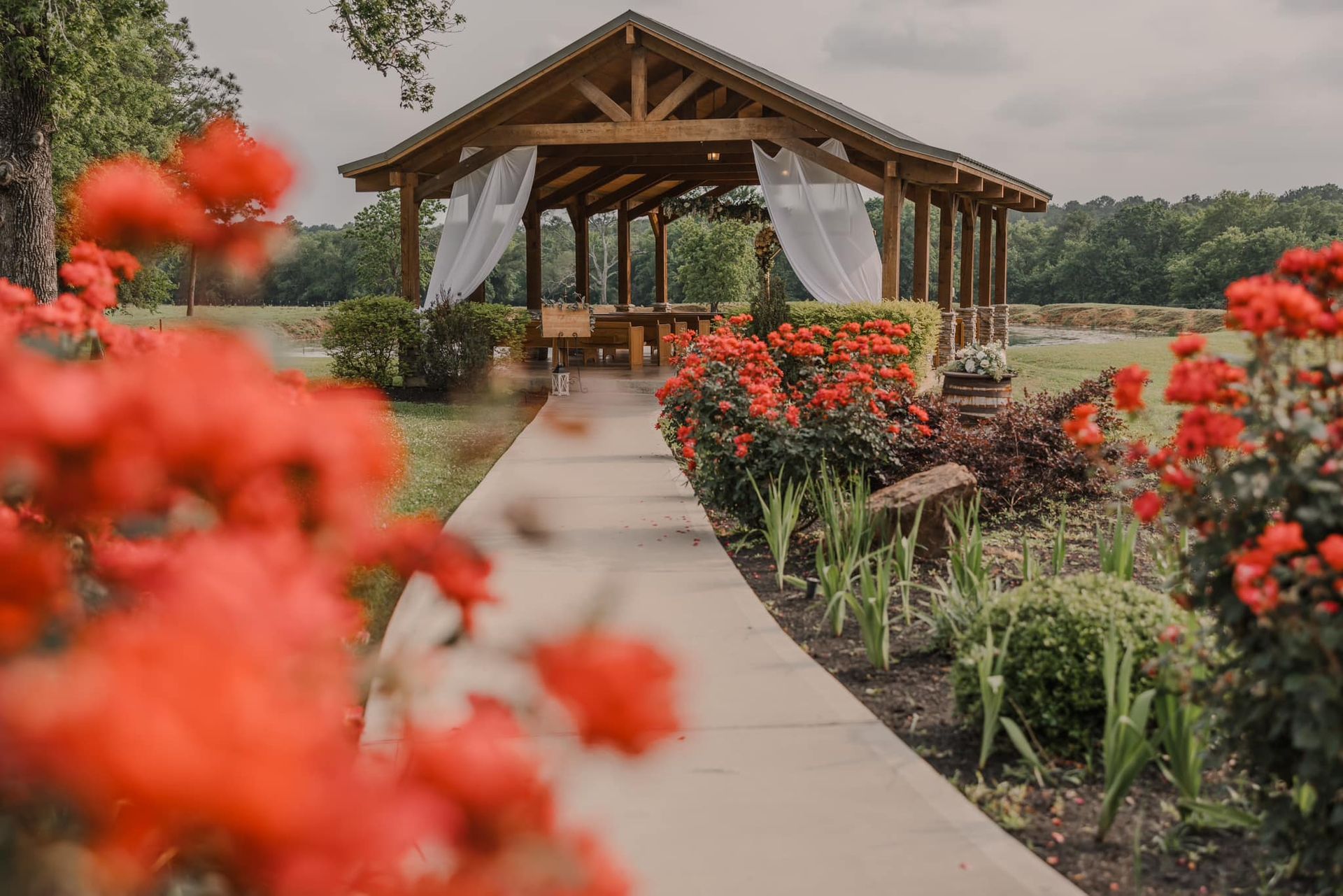 Garden walkway leading to a wooden pavilion, framed by red flowers and white drapes.