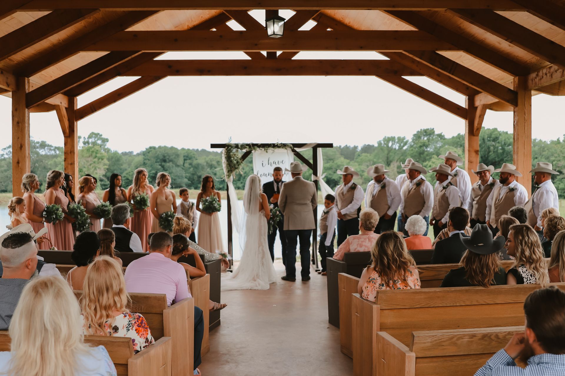 Outdoor wedding ceremony under a wooden pavilion with guests seated and a couple at the altar