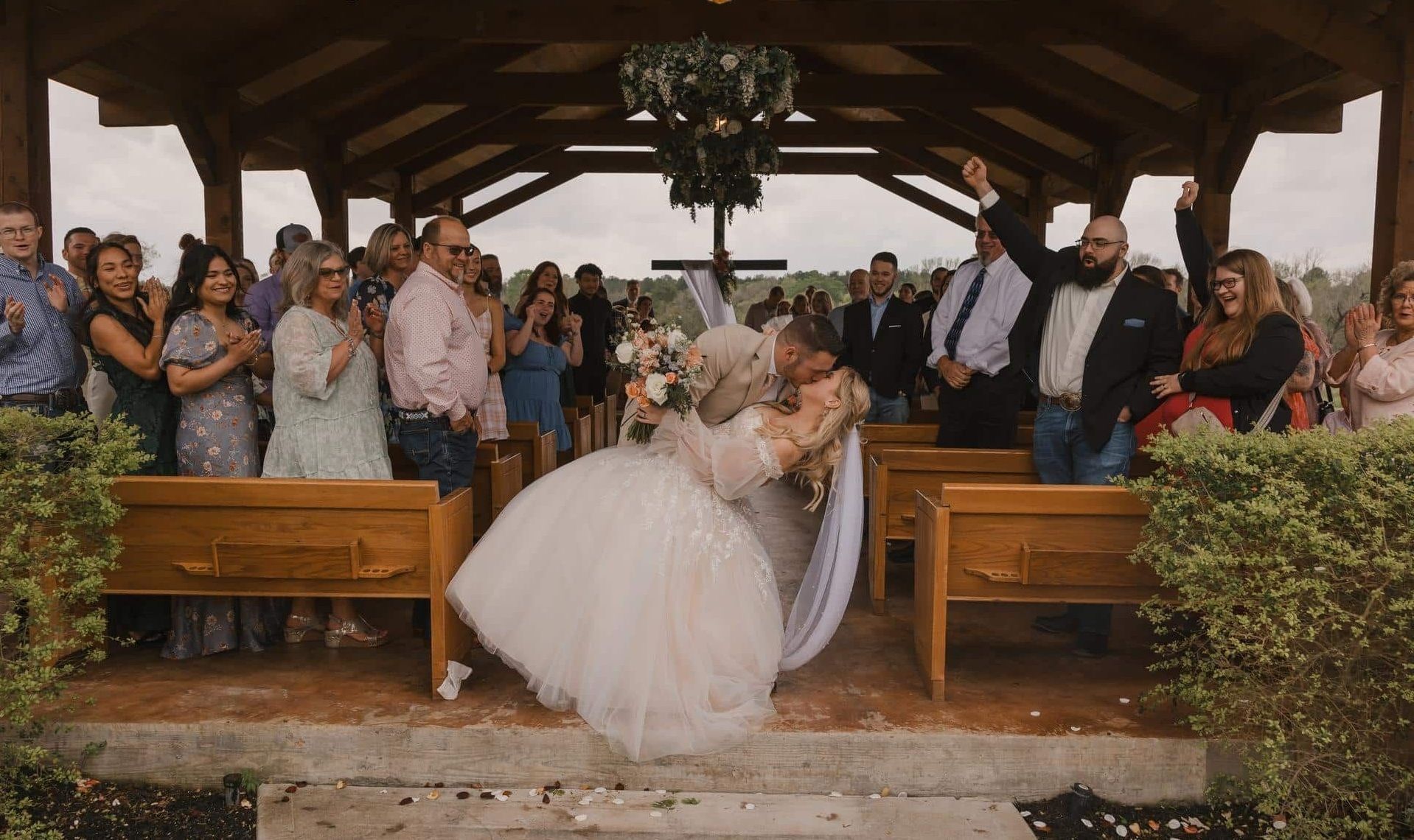 Bride and groom kiss at the altar during an outdoor wedding ceremony, with guests standing behind them.