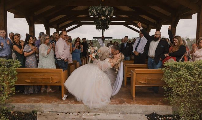 Bride and groom kiss at the altar during an outdoor wedding ceremony, with guests standing behind them.