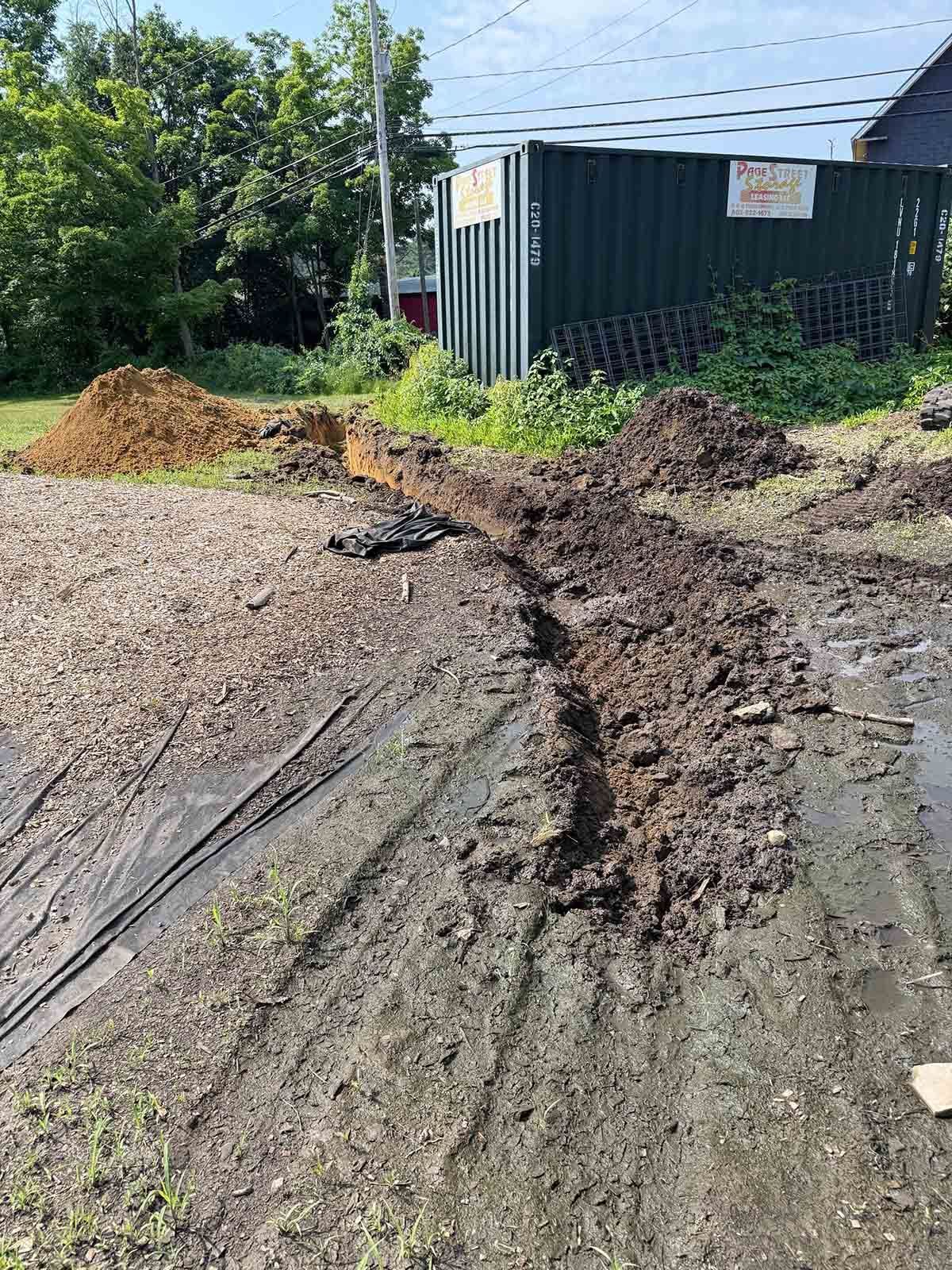 Mud-filled dirt road beside a green shipping container and piles of soil under bright sun