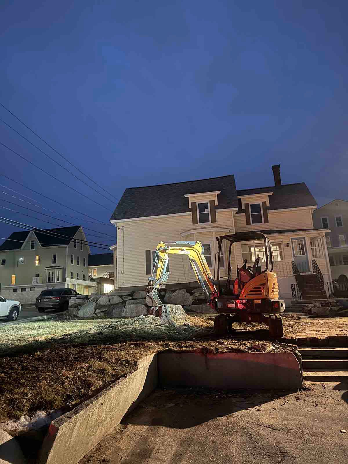 Excavator working in front of a house at dusk, with a bulldozer blade and debris in the yard.