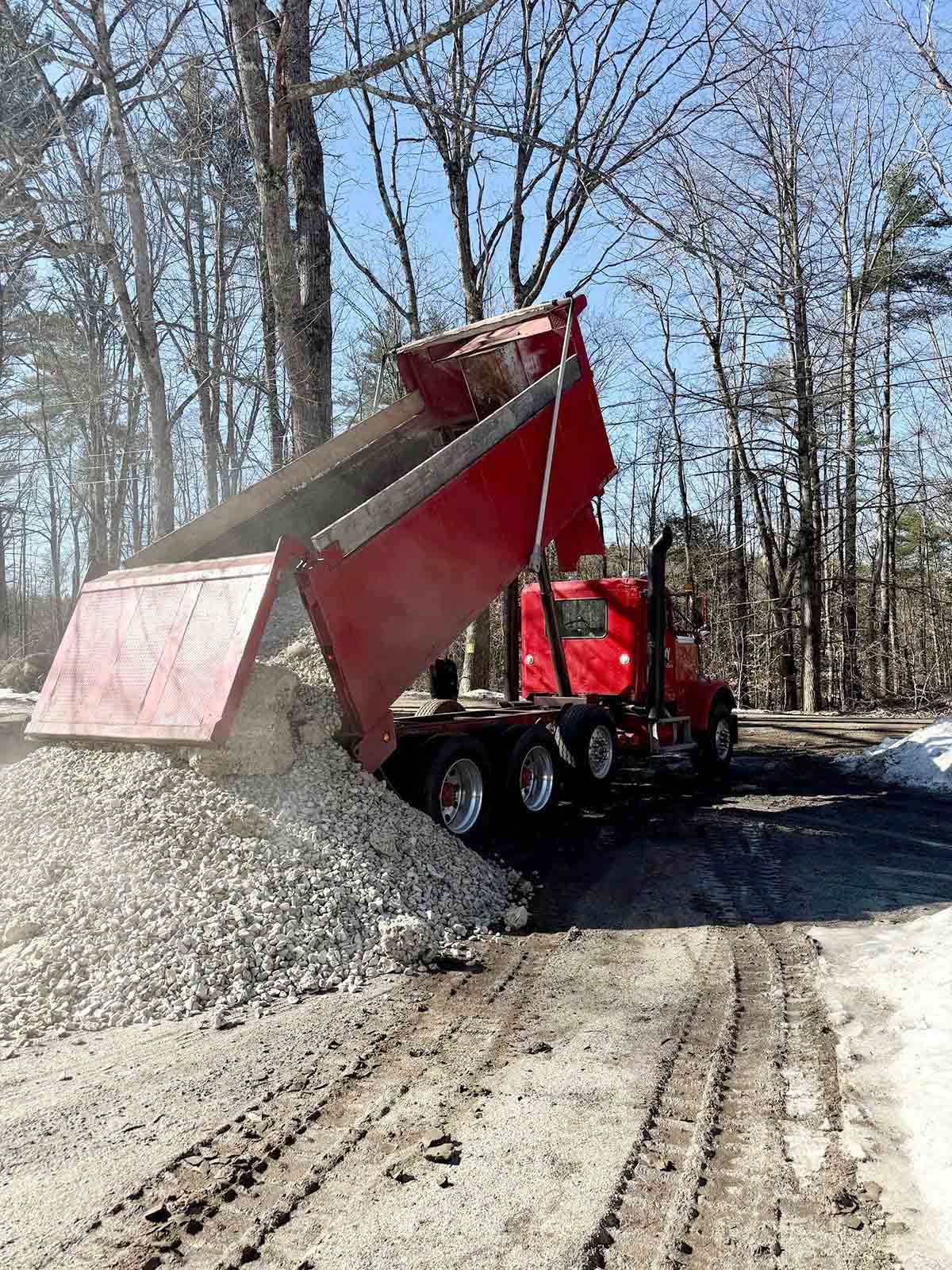 Red dump truck unloading gray gravel onto a dirt road in a wooded area