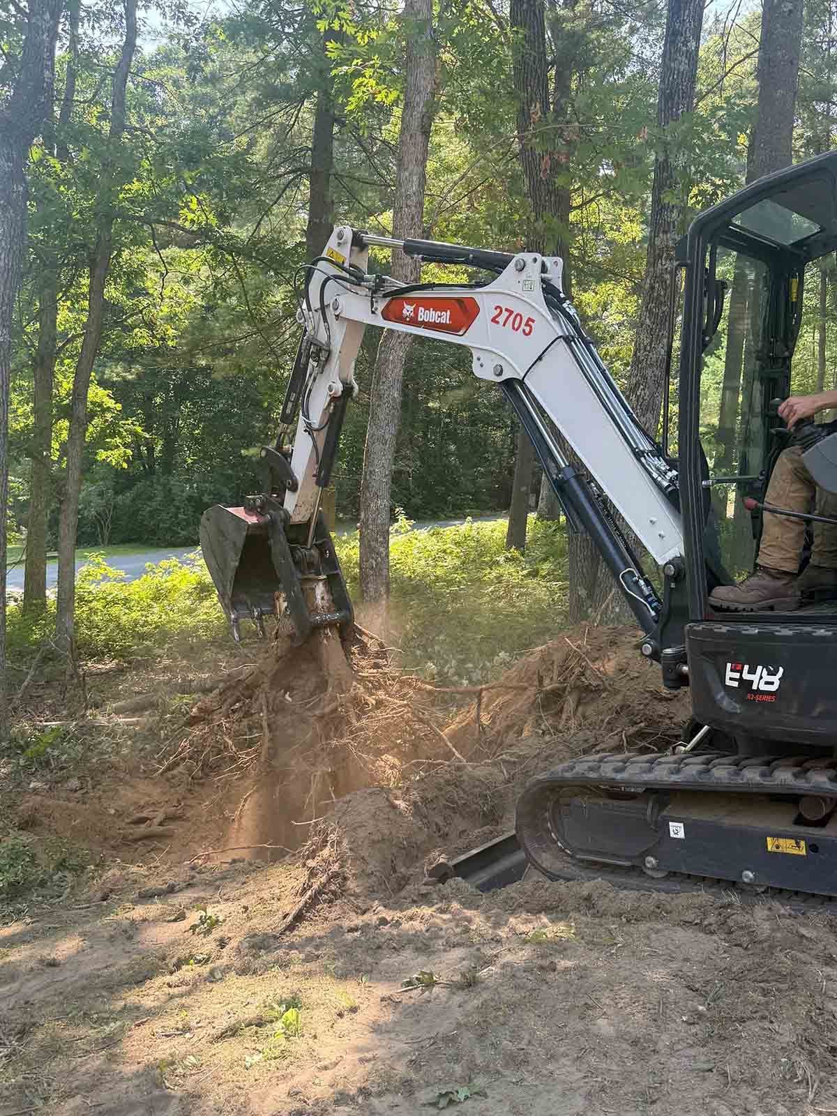 Excavator digging dirt in a wooded area, with a person operating it in the cab.