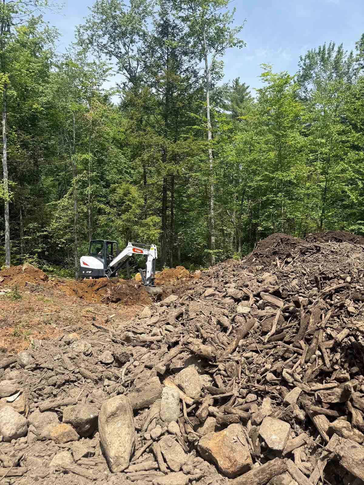 Excavator working beside a large rock pile in a wooded clearing under green trees