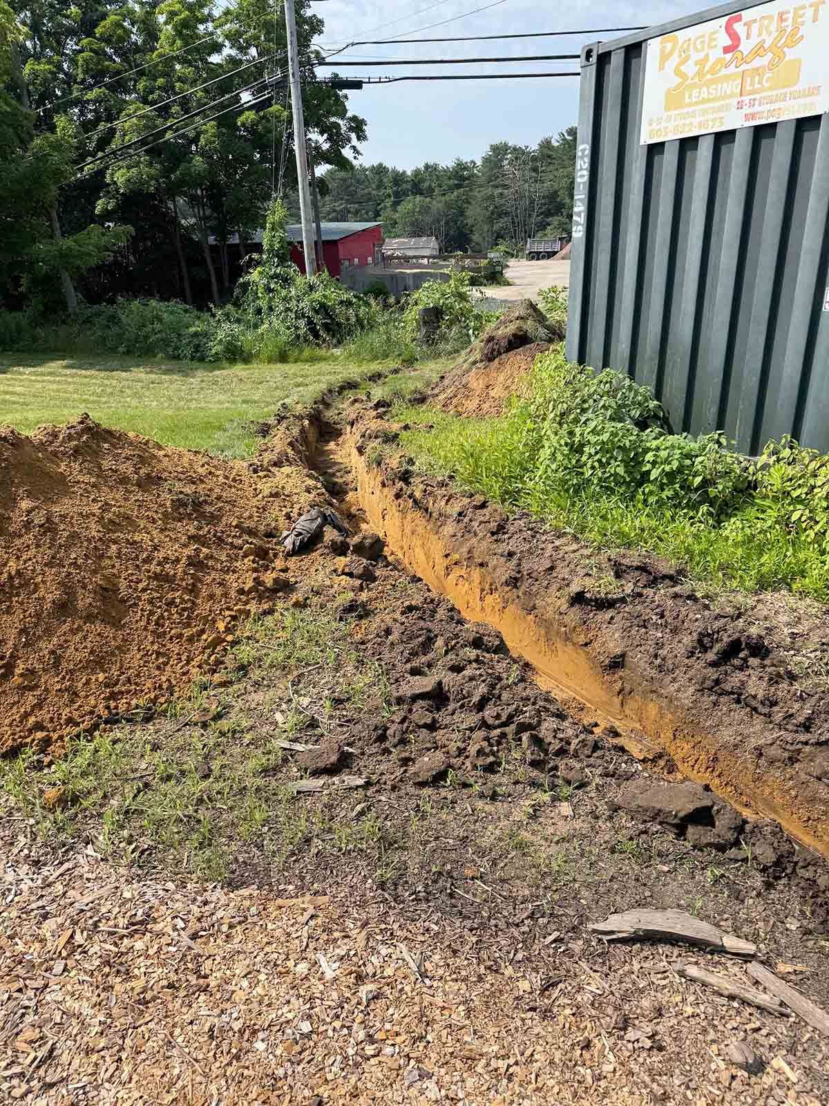 Mud-filled trench along a gravel path beside a metal building and grassy roadside.