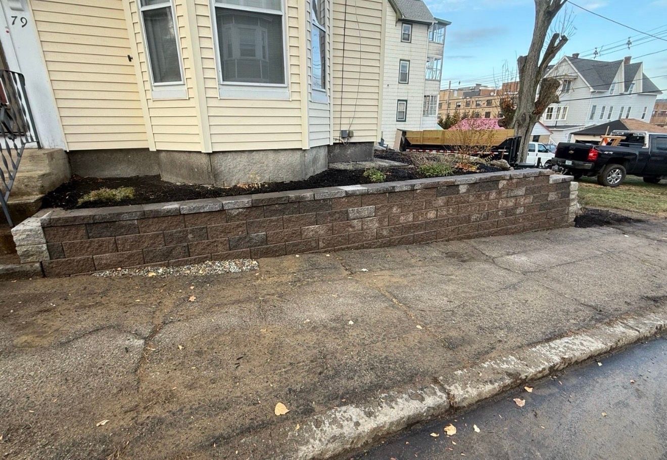Damaged brick retaining wall and uneven sidewalk beside a yellow house and street.